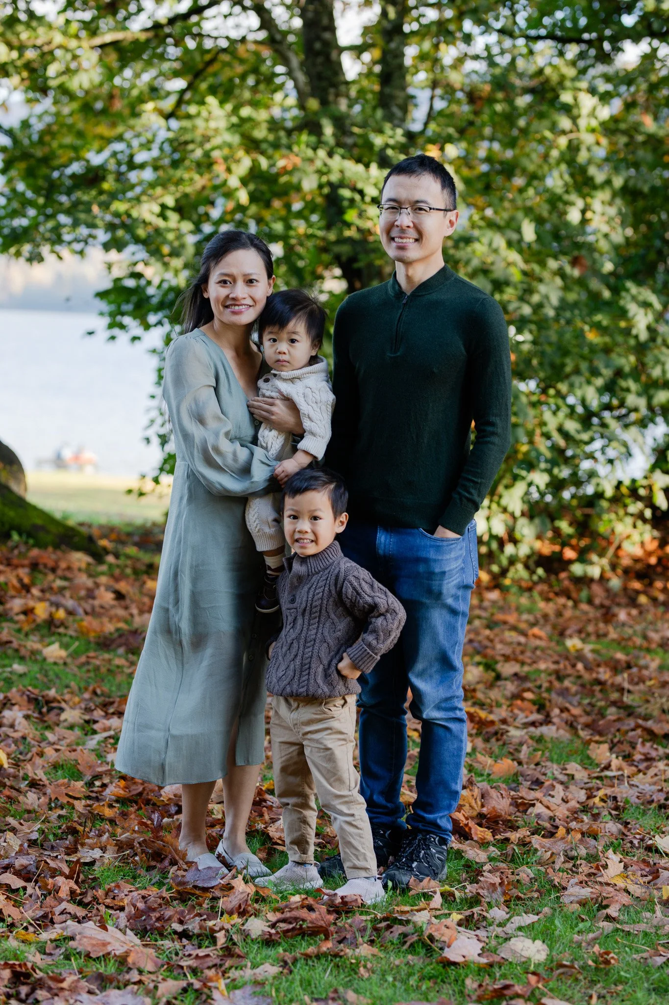 Family portrait of four standing outdoors in autumn leaves, with trees and a lake in the background.