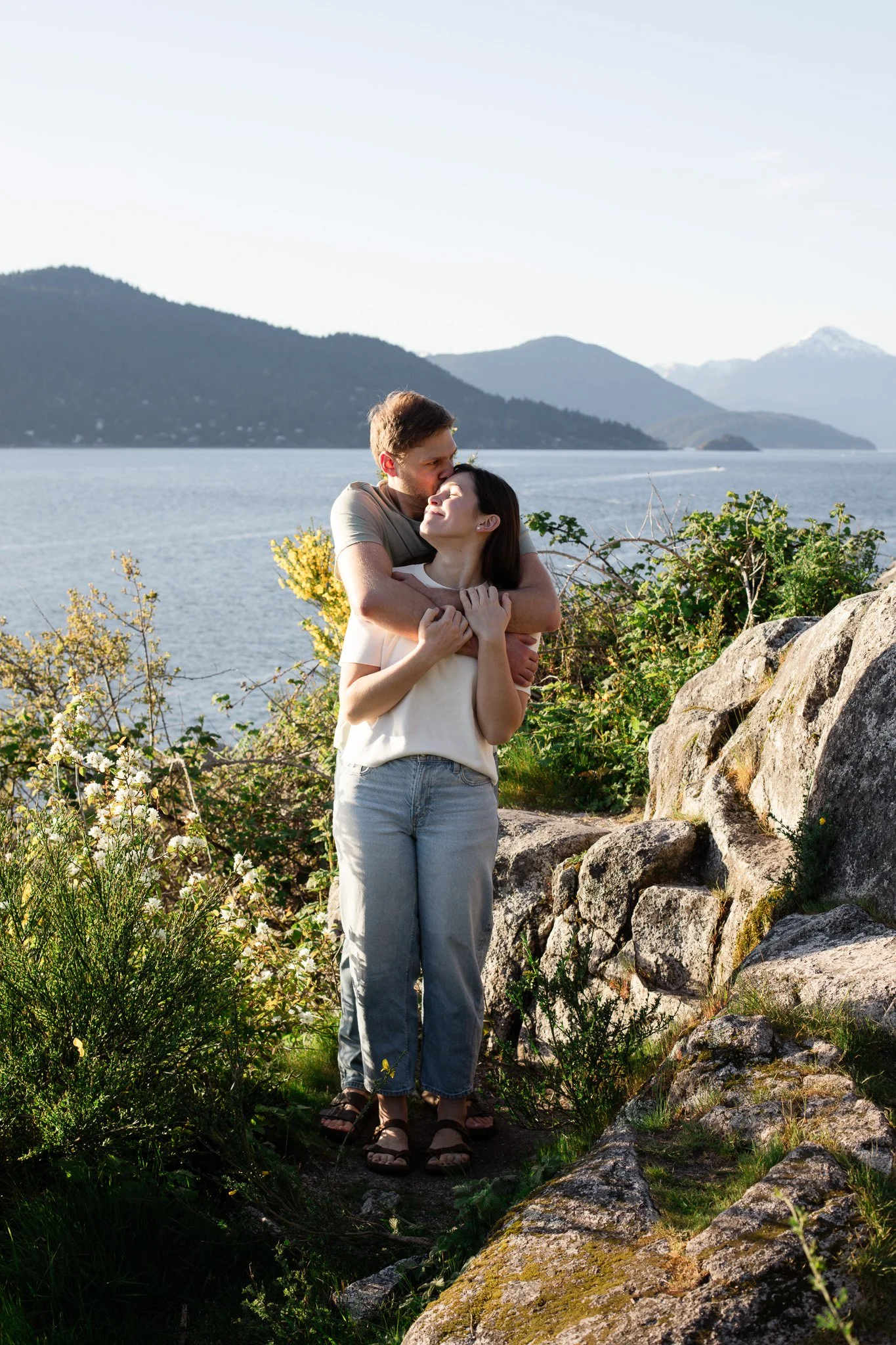 A couple embracing outdoors near a body of water with mountains in the background.