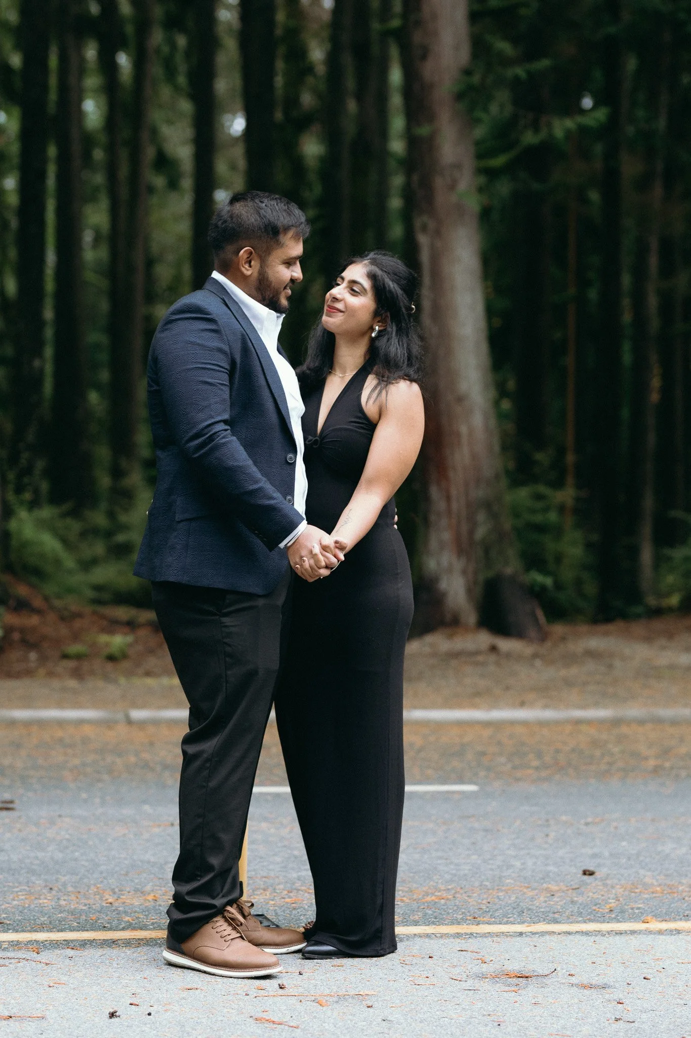 A couple holding hands and gazing at each other in a forest area, dressed in formal attire.