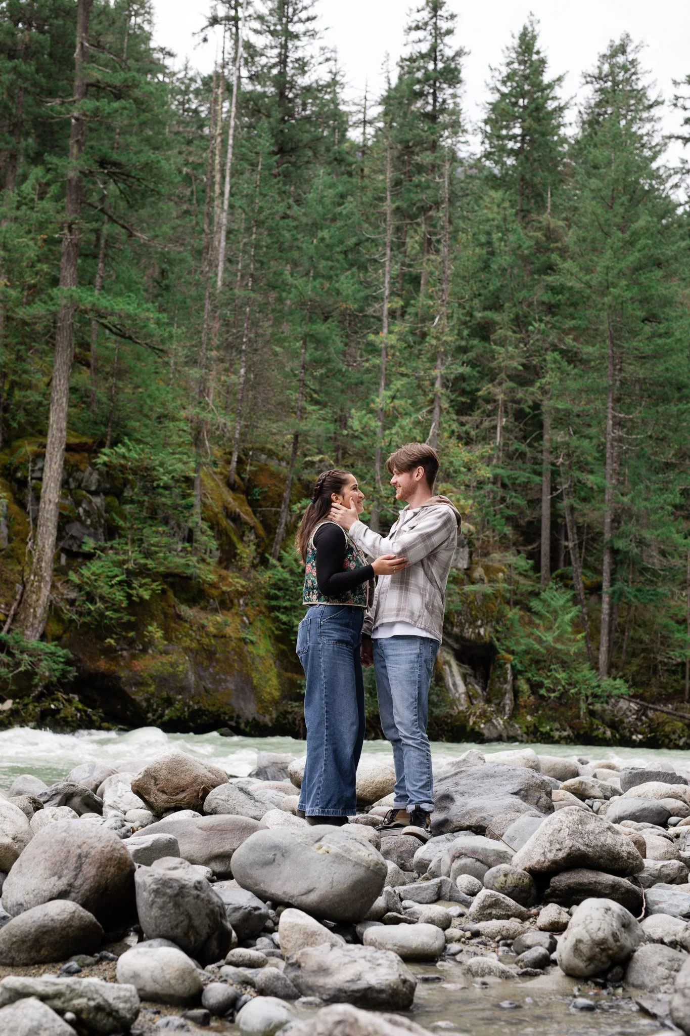 A young couple stands on rocks in a river, surrounded by a forest of tall green trees, as they look lovingly at each other.