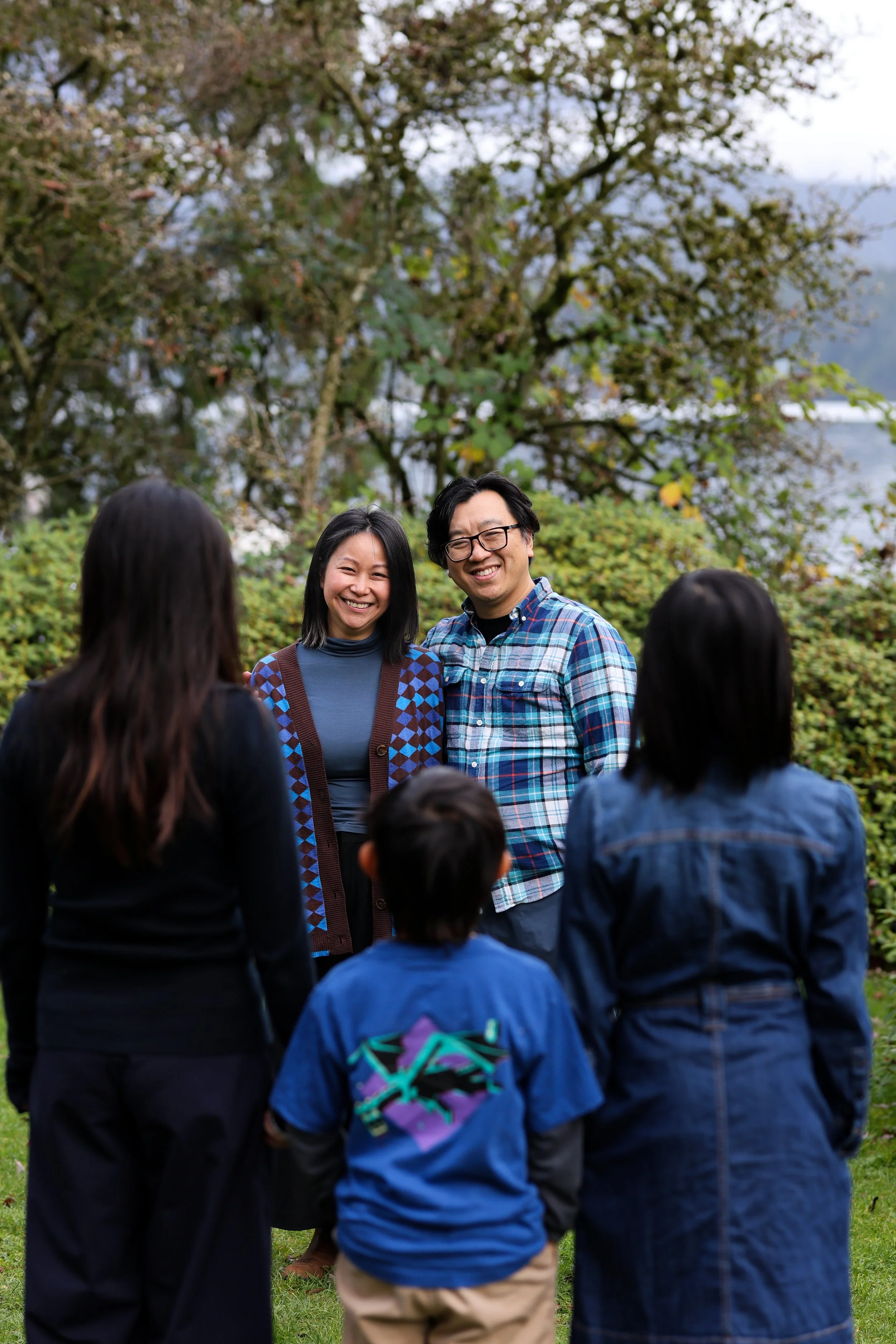 A group of six people outdoor, facing each other and smiling, with trees and bushes in the background.