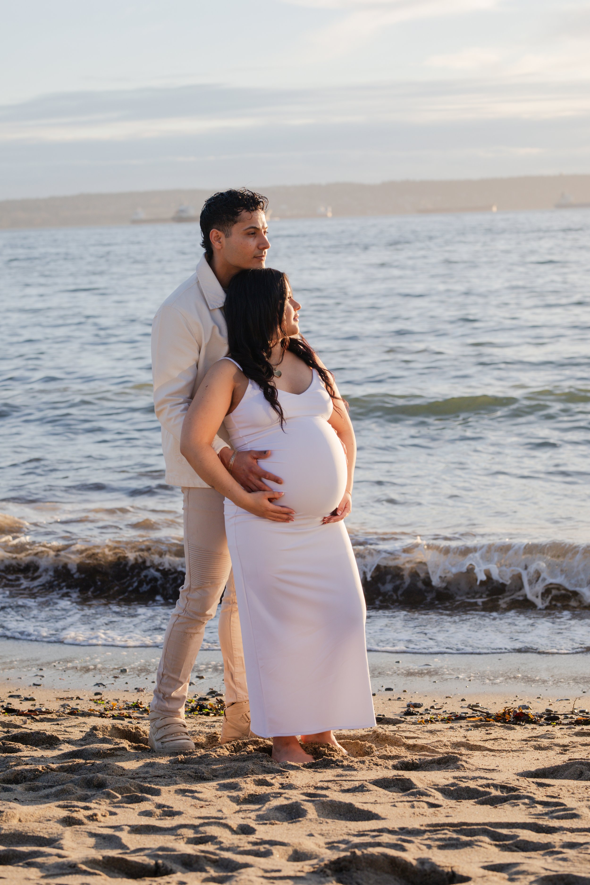 A pregnant woman and a man standing on the beach near the water, with a car and ships visible in the background.