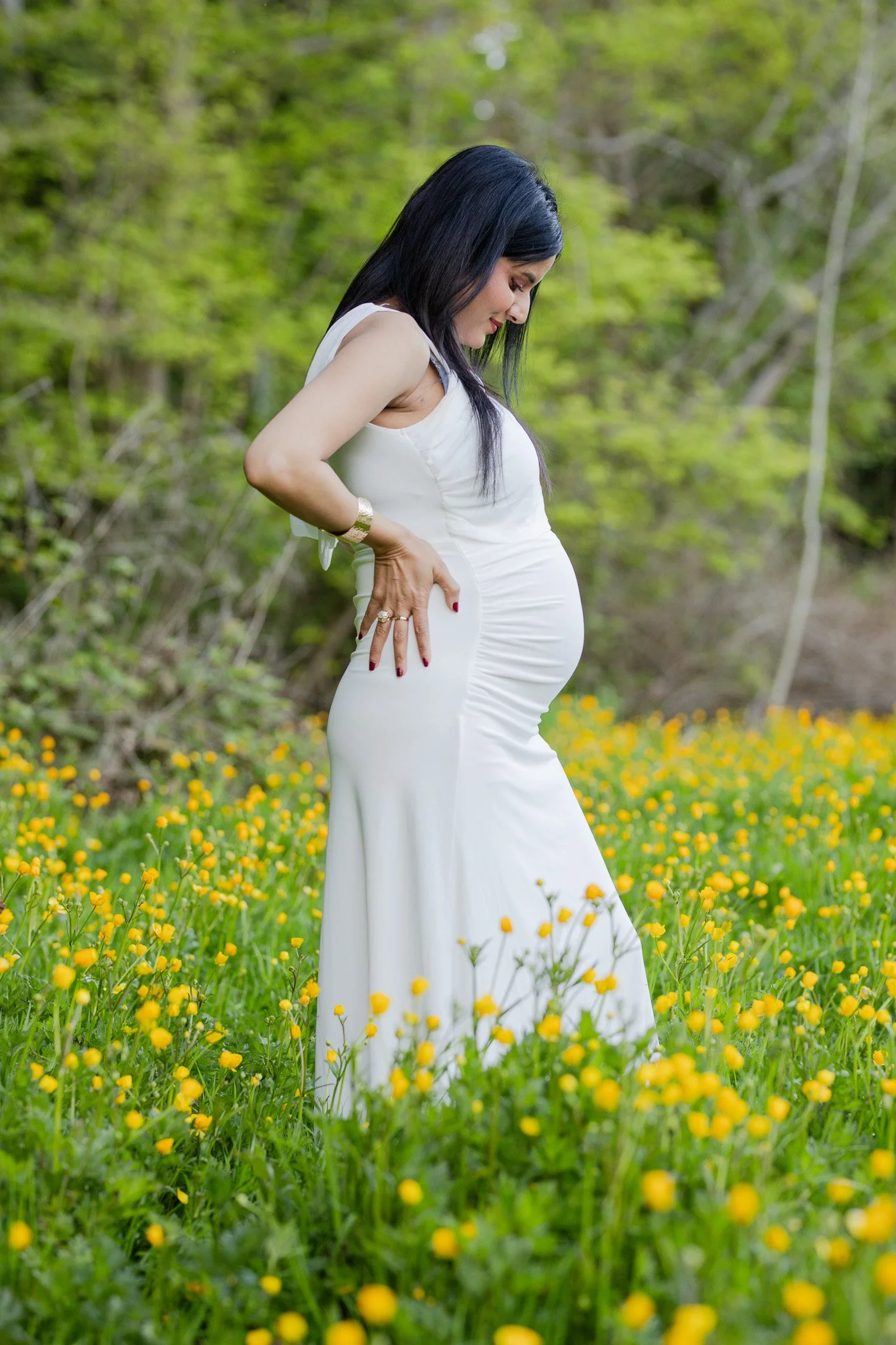 A pregnant woman in a white dress standing in a field of yellow flowers with green trees in the background, looking down at her belly.