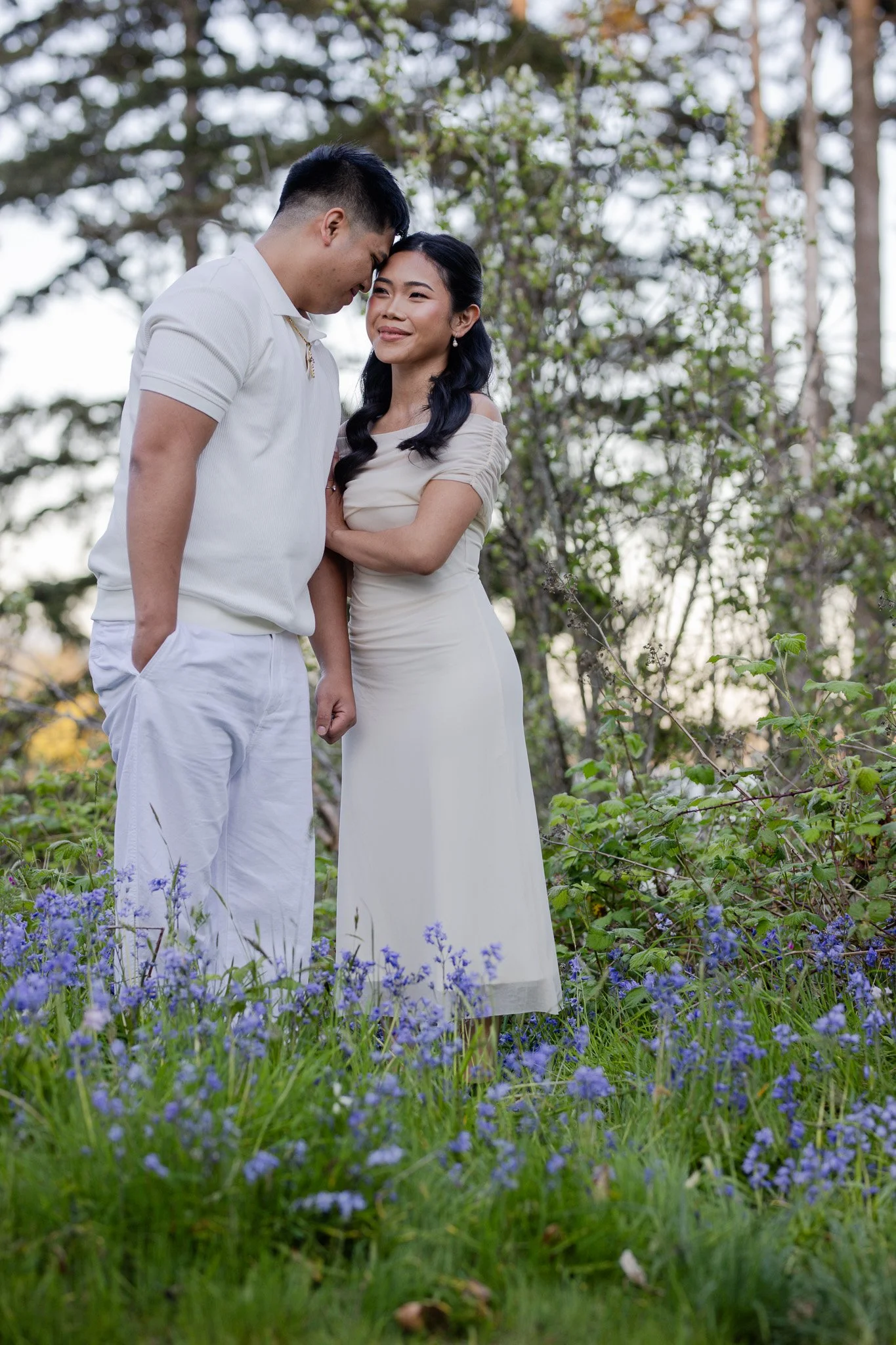 A young couple stands close together in a field of purple violets, with trees and cloudy sky in the background. They are smiling softly, with the man's forehead touching the woman's forehead.