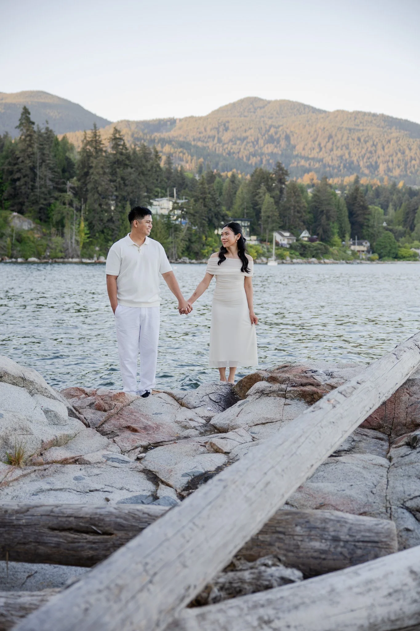 A couple holding hands standing on rocks by a body of water with trees and mountains in the background.