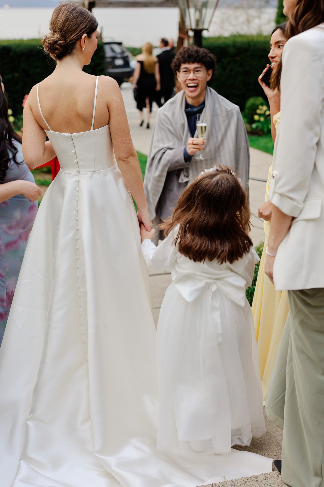 Group of women and children dressed in formal attire, including a bride in a white wedding gown, standing outdoors on a sidewalk during a celebration.