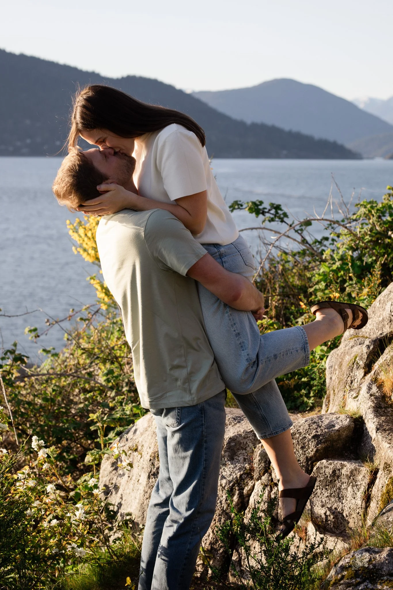 A man is lifting a woman while they kiss near a lake with mountains in the background.