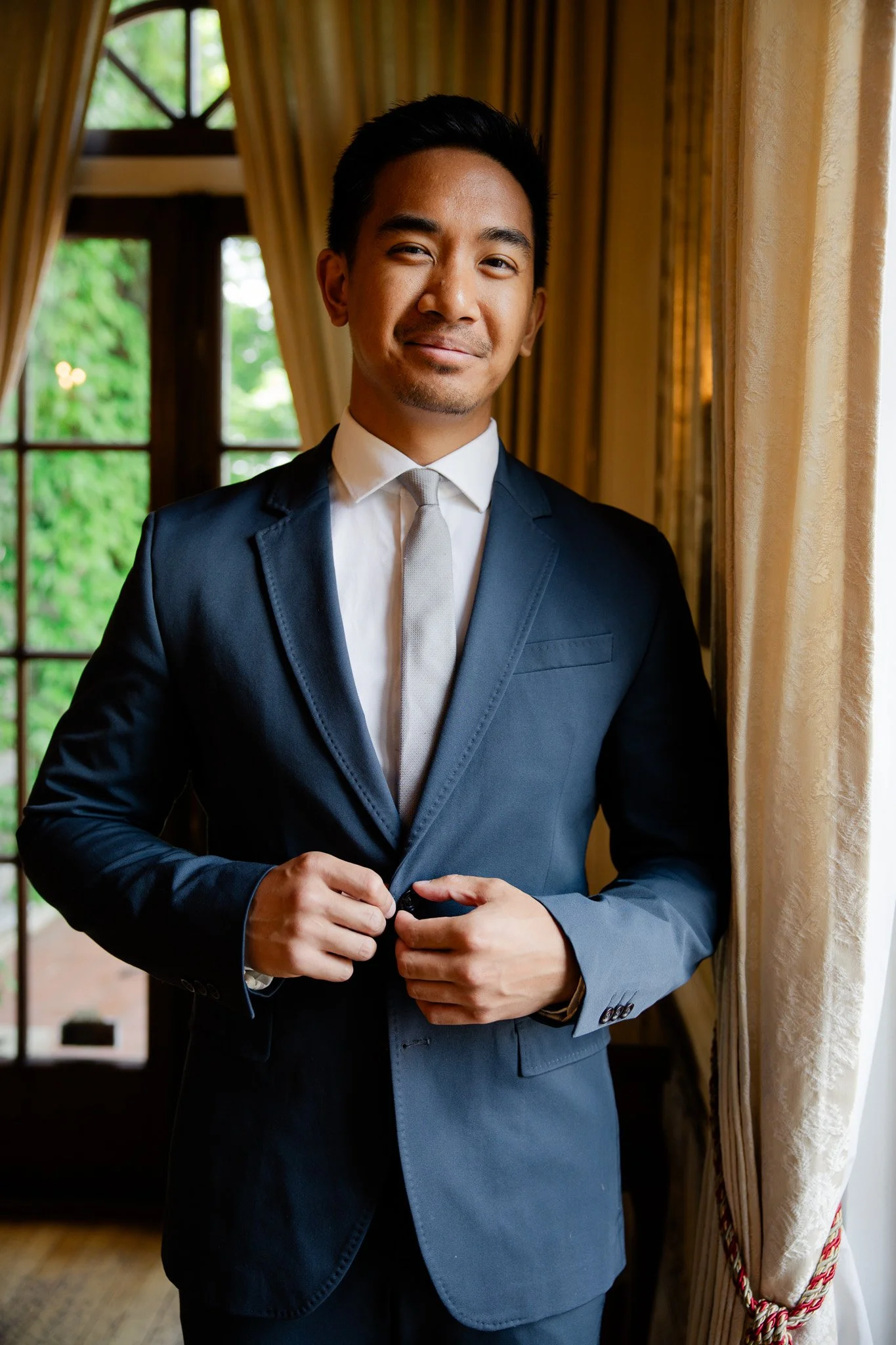 A young man in a navy suit and white shirt, standing indoors near a window with yellow curtains, smiling softly at the camera.