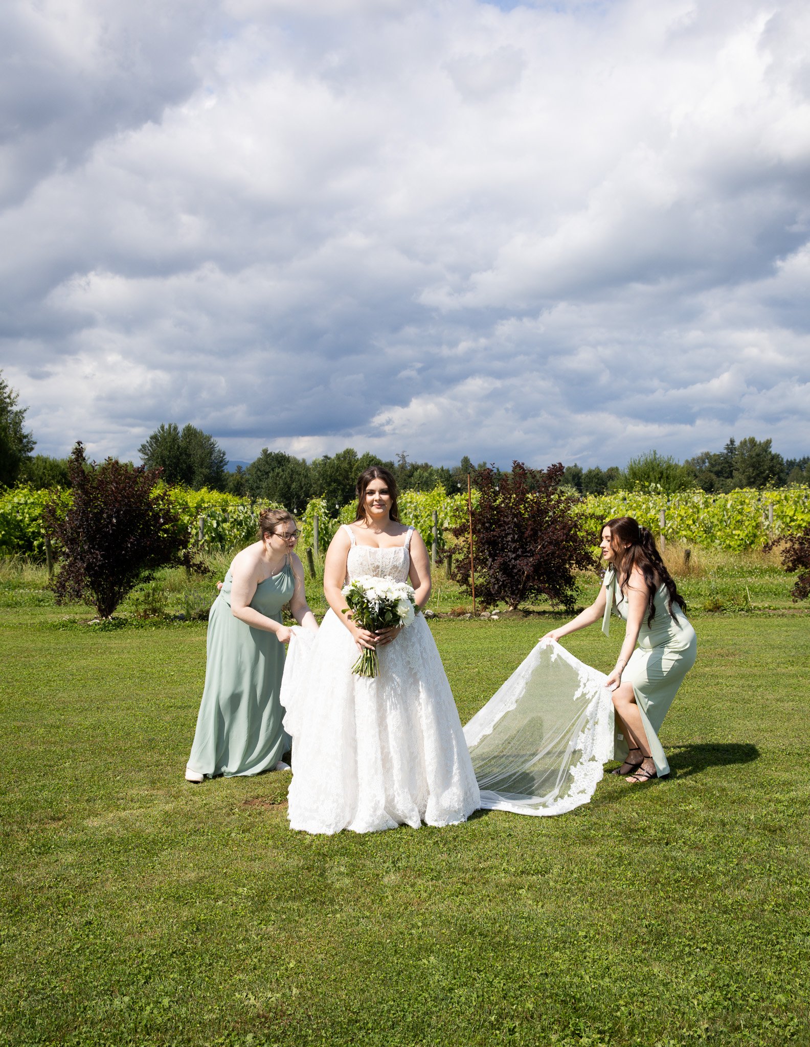 A bride in a white wedding dress holding a bouquet of white flowers, being assisted by three women in green dresses during an outdoor wedding ceremony on a grassy field, with trees and a cloudy sky in the background.