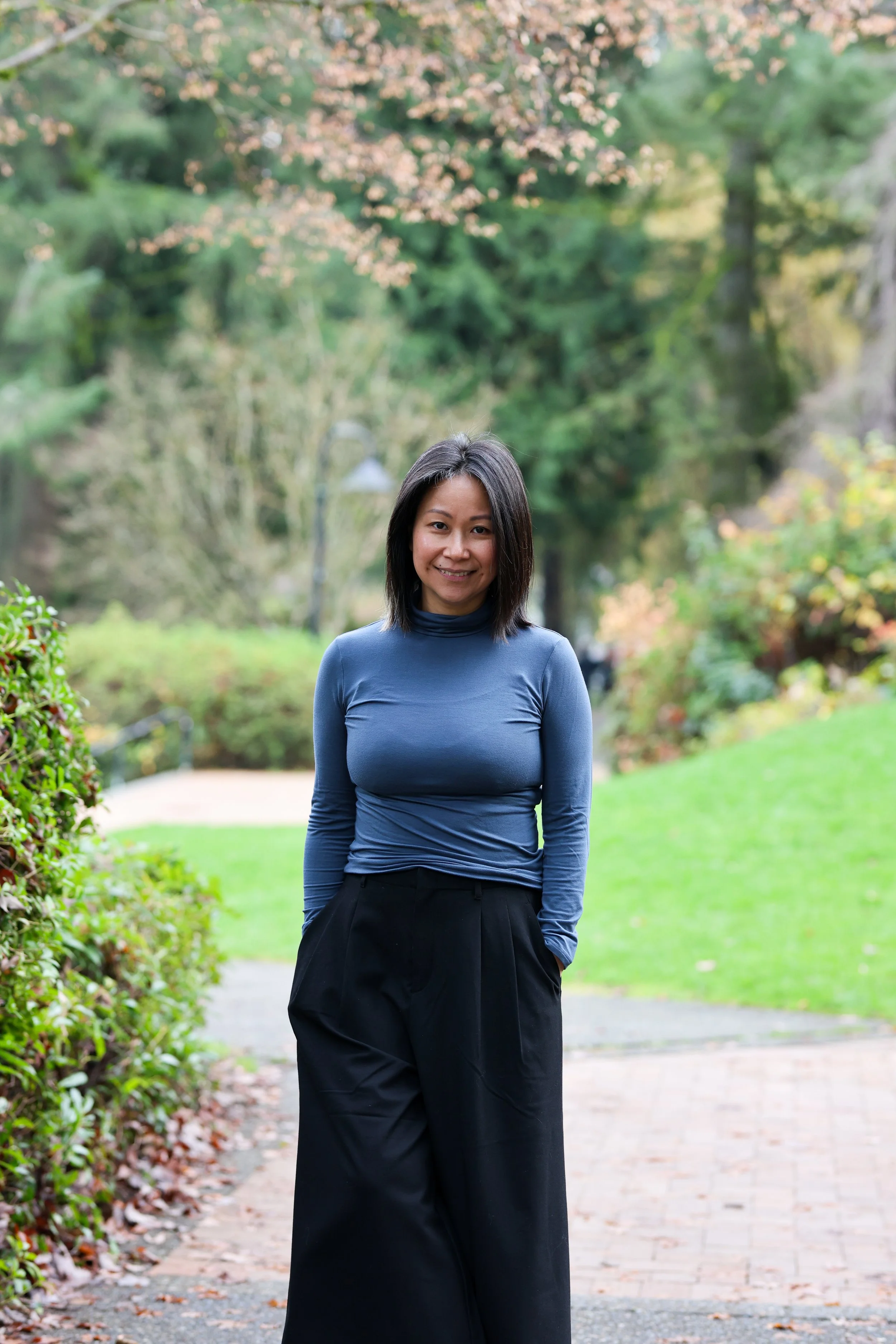 A woman standing on a park trail surrounded by greenery and trees, wearing a blue long-sleeve shirt and black wide-leg pants, smiling at the camera.