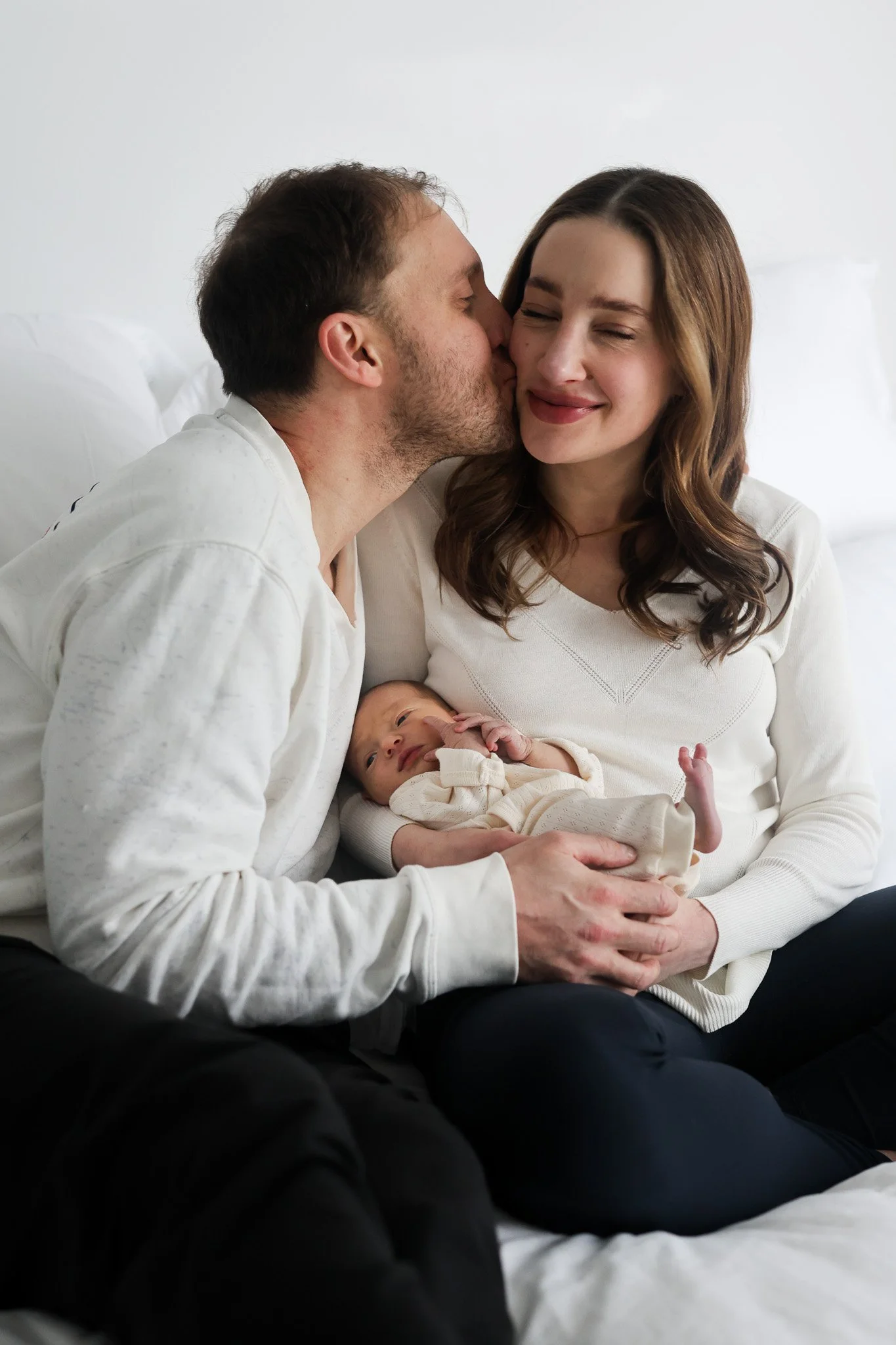 A man kisses a woman on the cheek, who is holding a newborn baby in her arms. The scene appears to be a family moment in a bright, cozy room with a white background.