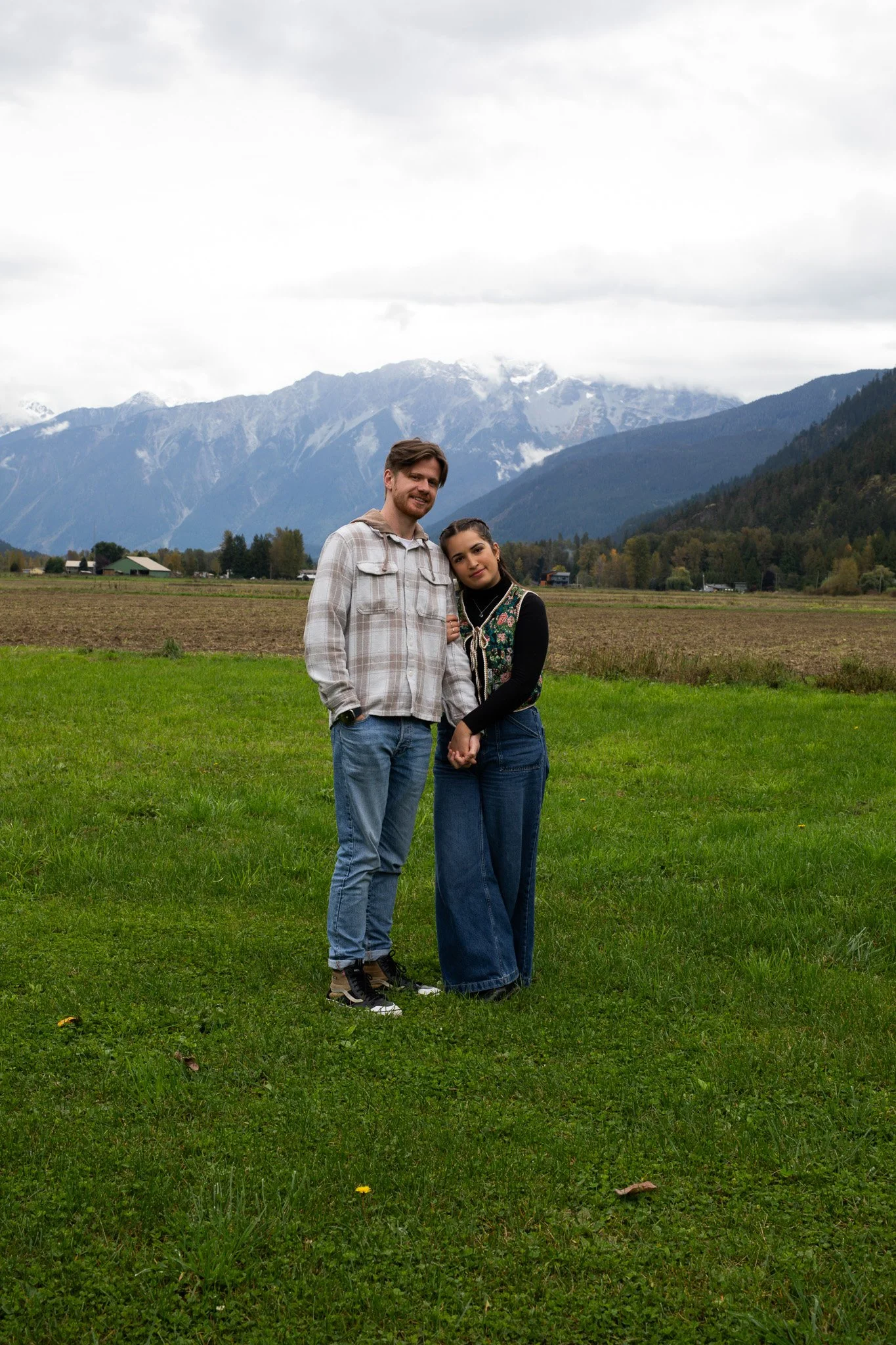 A young man and woman stand together on a grassy field with mountains and cloudy sky in the background.