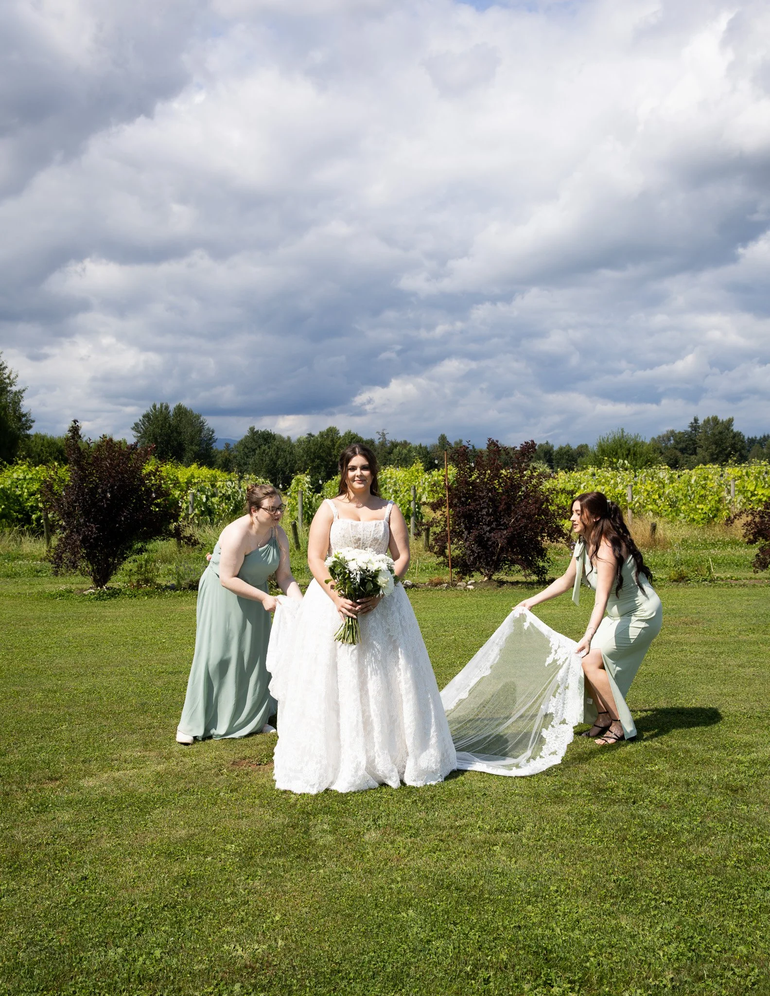 A bride in a white wedding dress holding a bouquet of white flowers stands on a grassy field with four bridesmaids around her, two of whom are holding a wedding veil. There are trees and bushes in the background under a cloudy sky.