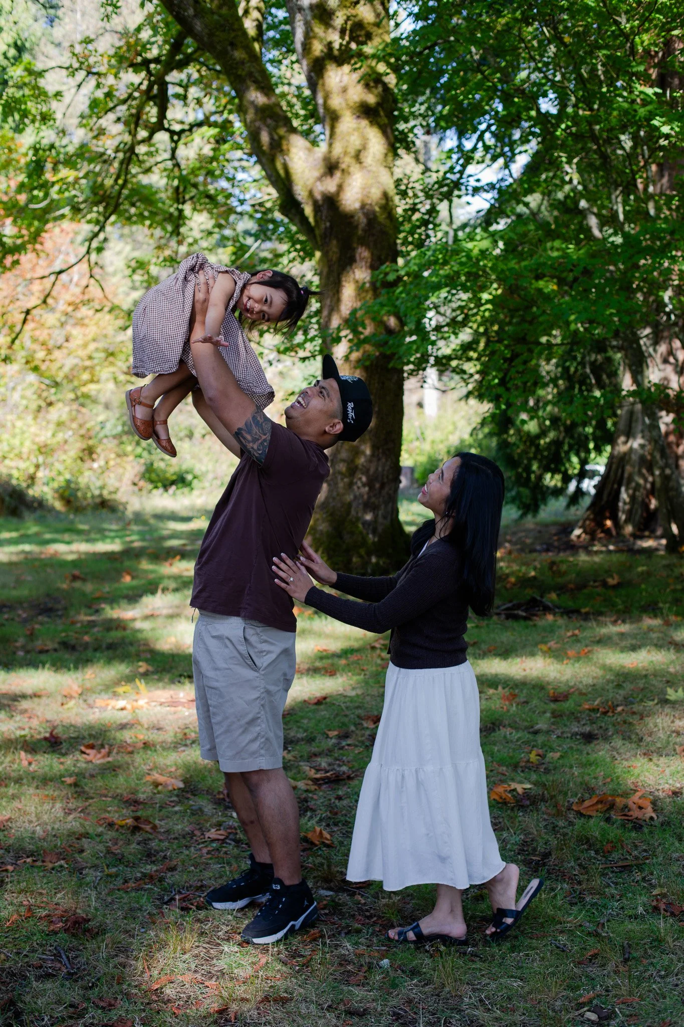 A man raising a young girl in a park, with a woman standing nearby smiling, surrounded by trees and greenery.