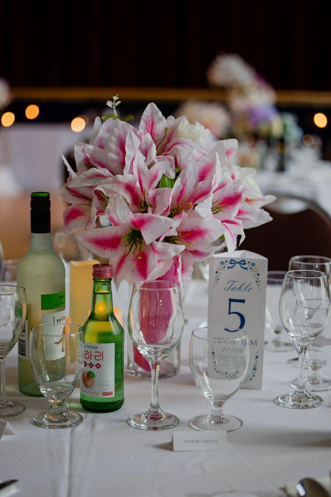 Close-up of a floral centerpiece with pink and white lilies on a table at a formal event, with various glassware and bottles surrounding it.