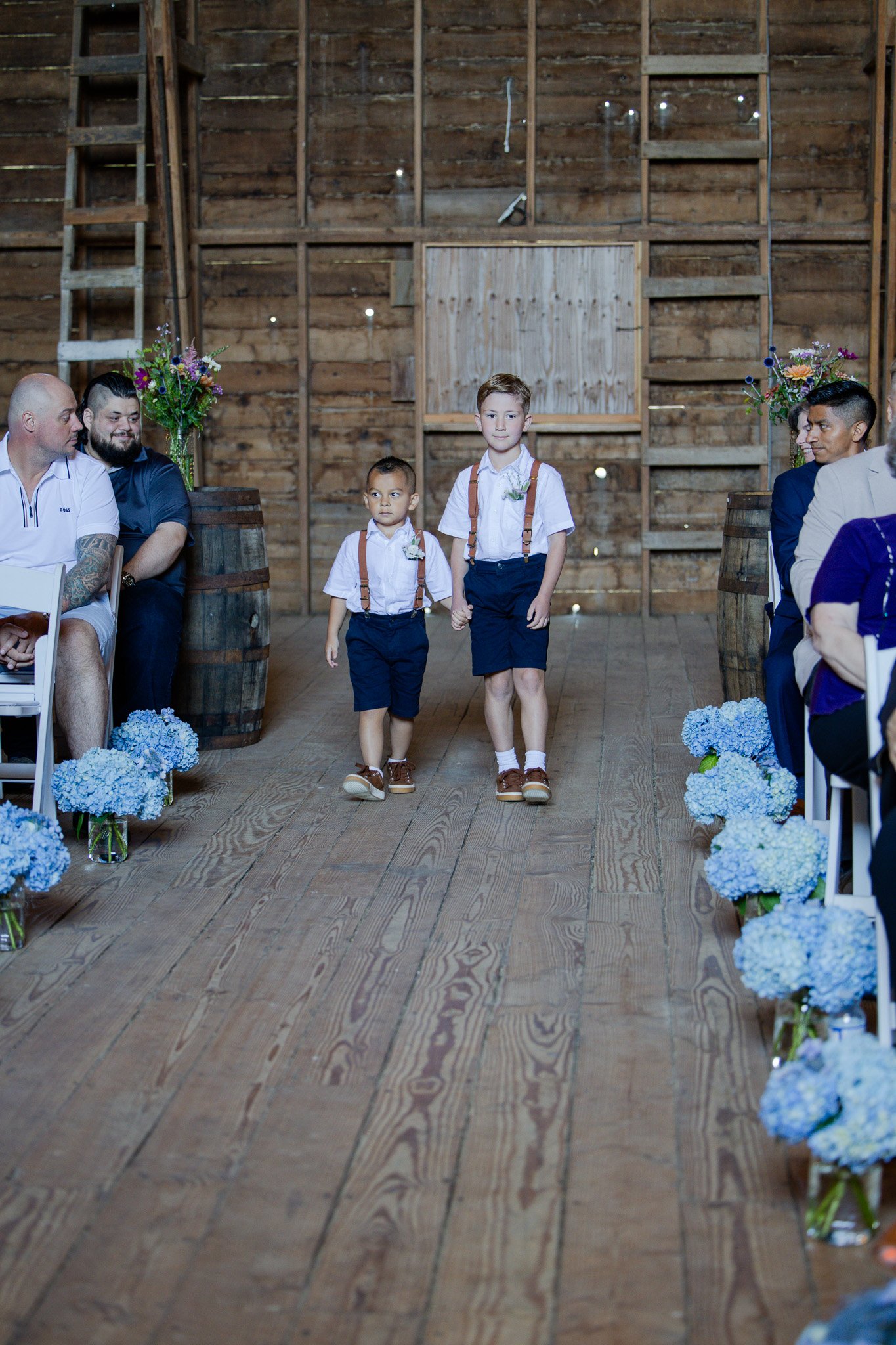 Two young boys dressed in white shirts, dark shorts, suspenders, and brown shoes walking hand-in-hand down the aisle of a wedding ceremony indoors with a rustic wooden wall in the background and floral arrangements on either side.