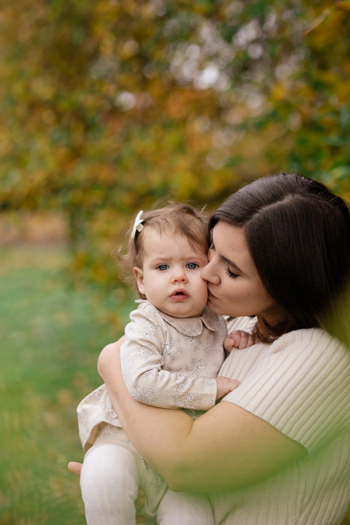 A woman holding a young girl outdoors during fall, with the woman kissing the girl on the cheek, and trees with yellow and green leaves in the background.