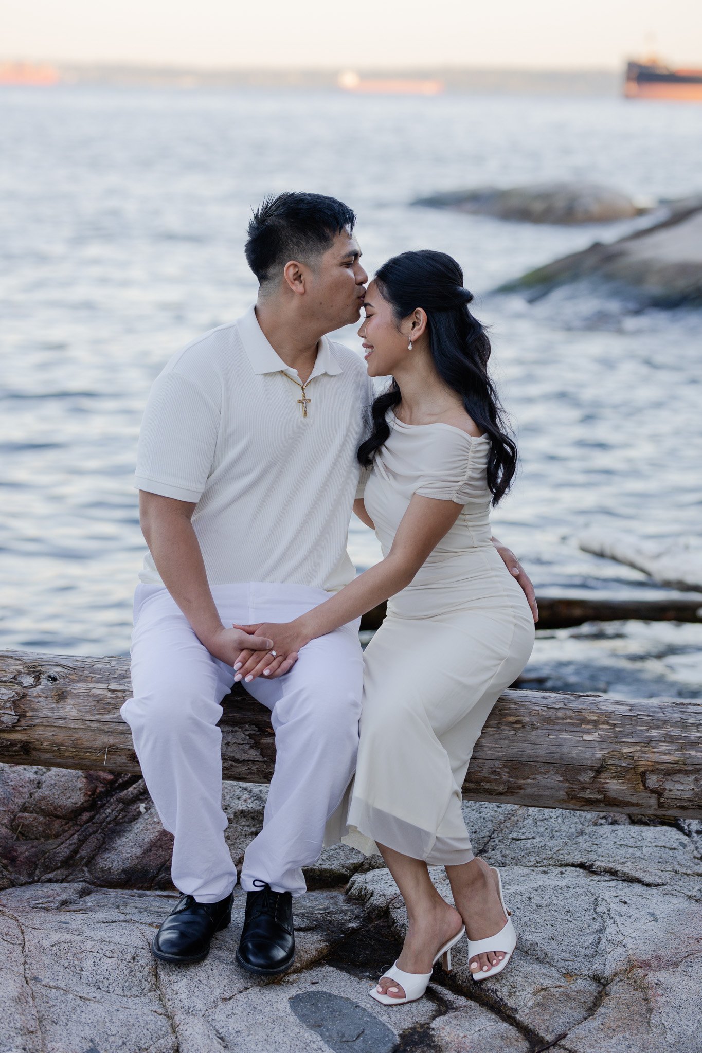 A couple sitting on a large log near the water, with the man kissing the woman's forehead. The woman is wearing a white dress and heels, and the man is dressed in white pants and a shirt with a cross necklace. There are ships in the background.