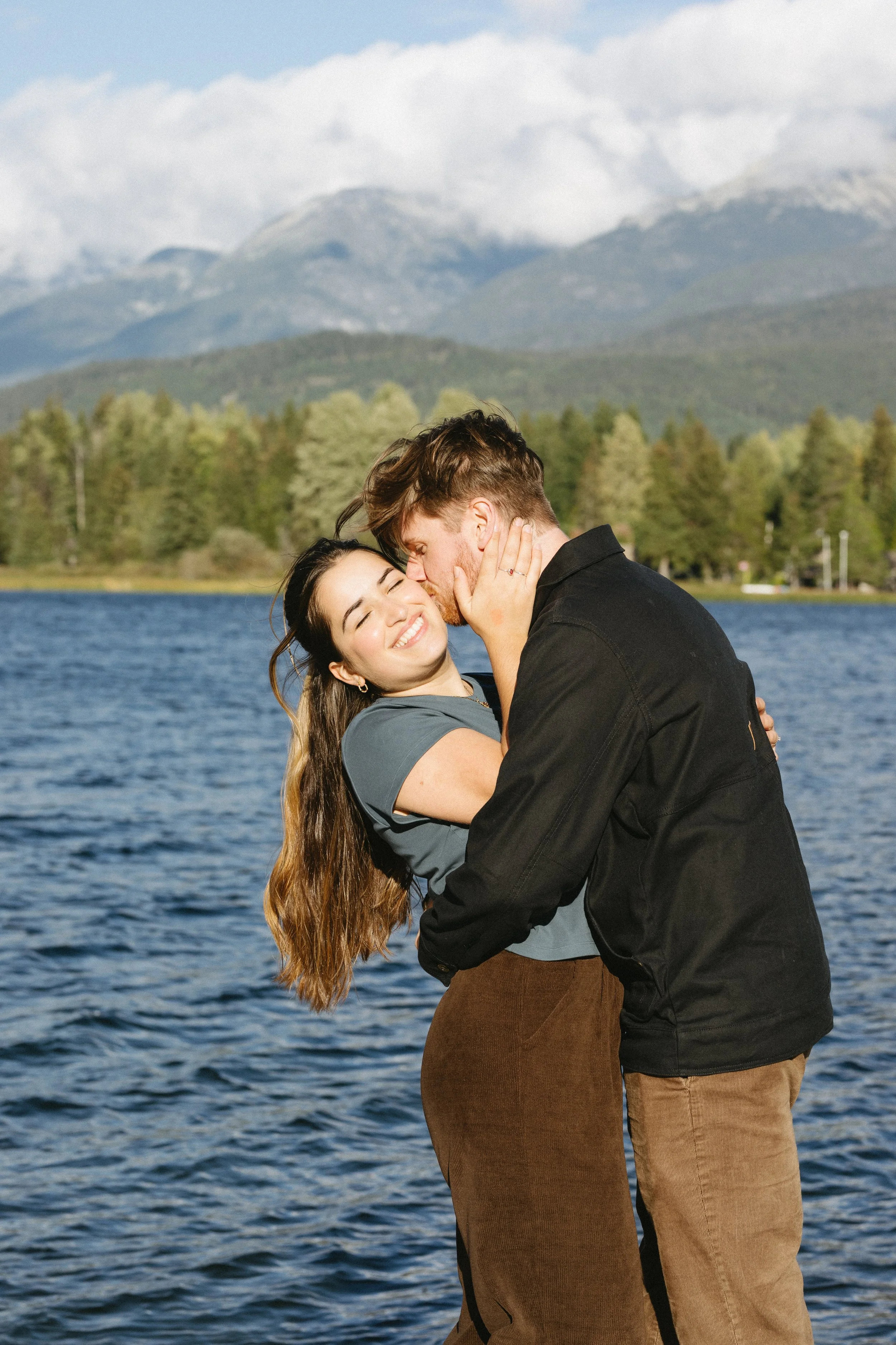 A couple happily embracing by a lake with mountains and trees in the background.