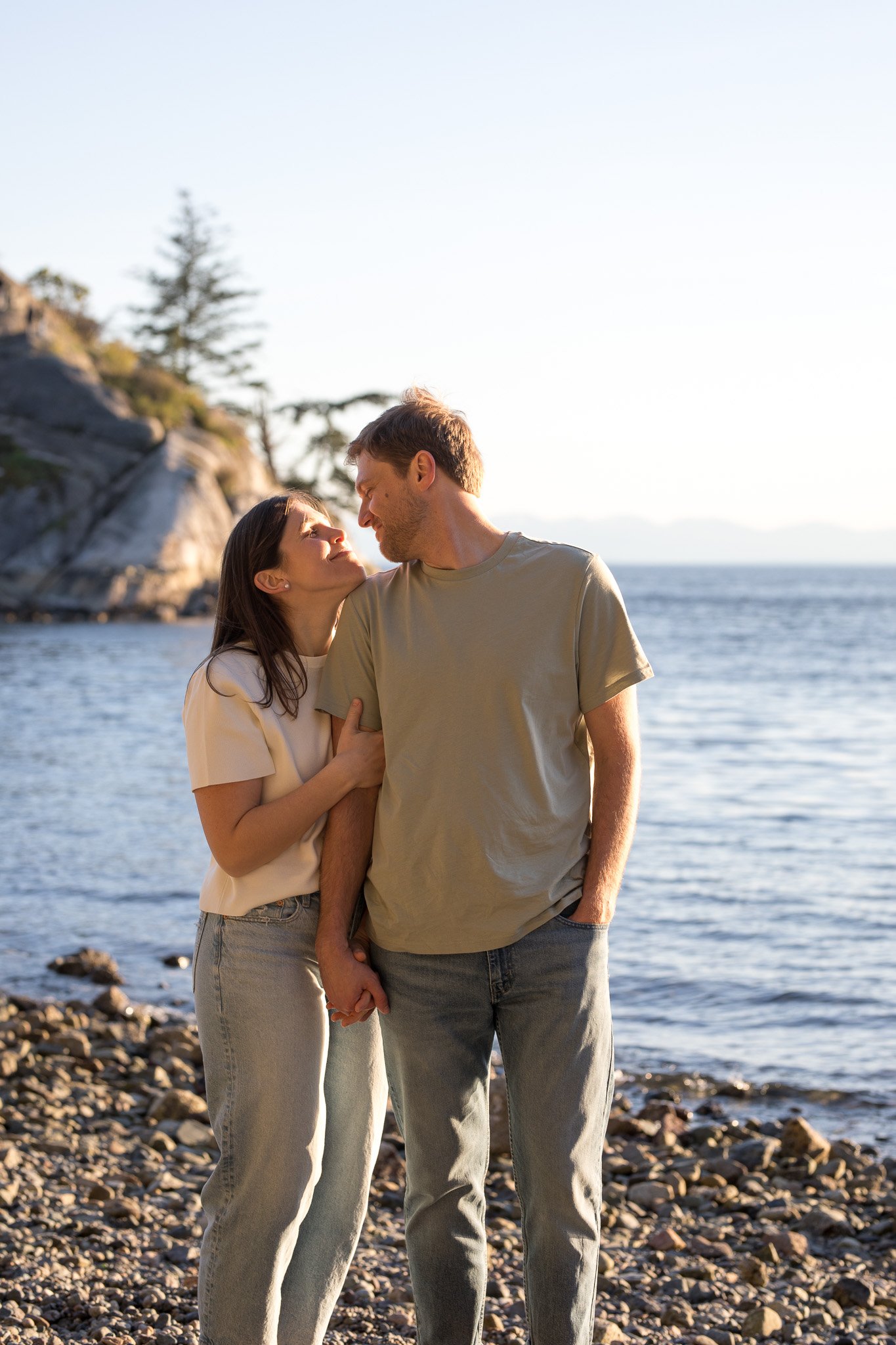 A couple standing on a rocky beach, gazing at each other, with water and trees in the background during sunset.