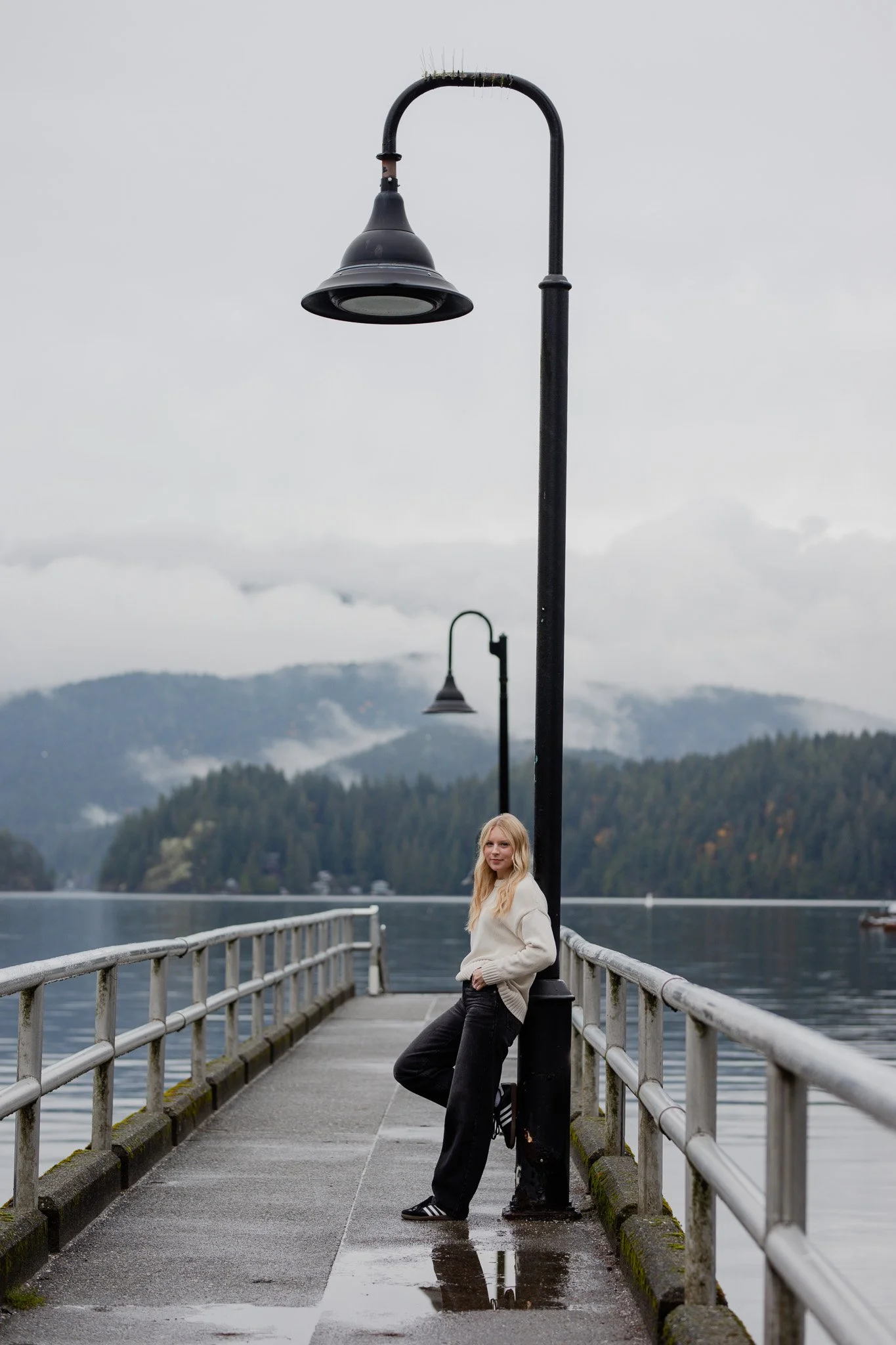 A young woman in a white sweater and black pants leaning against a light post on a pier by a lake, with mountains and cloudy skies in the background.