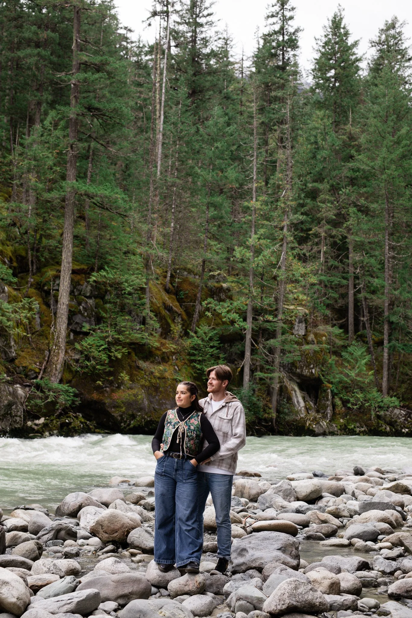 A couple standing on rocks by a flowing river, surrounded by a dense forest of tall green trees.