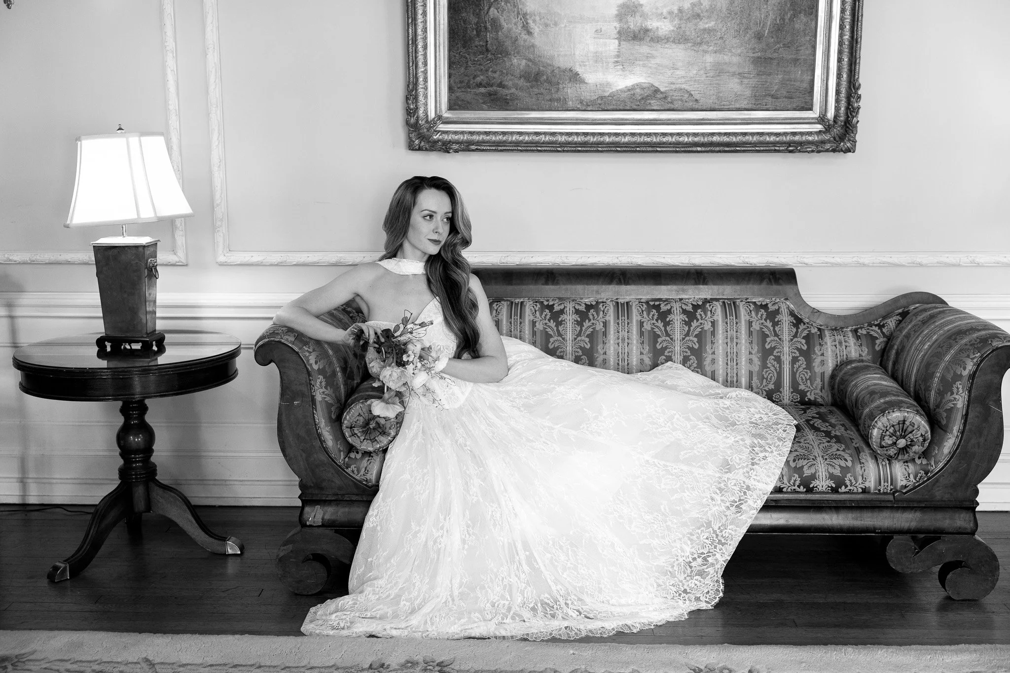 A woman in a wedding dress sitting on a vintage sofa in a decorated room with ornate wall trim, a large framed painting, and a lamp on a side table.