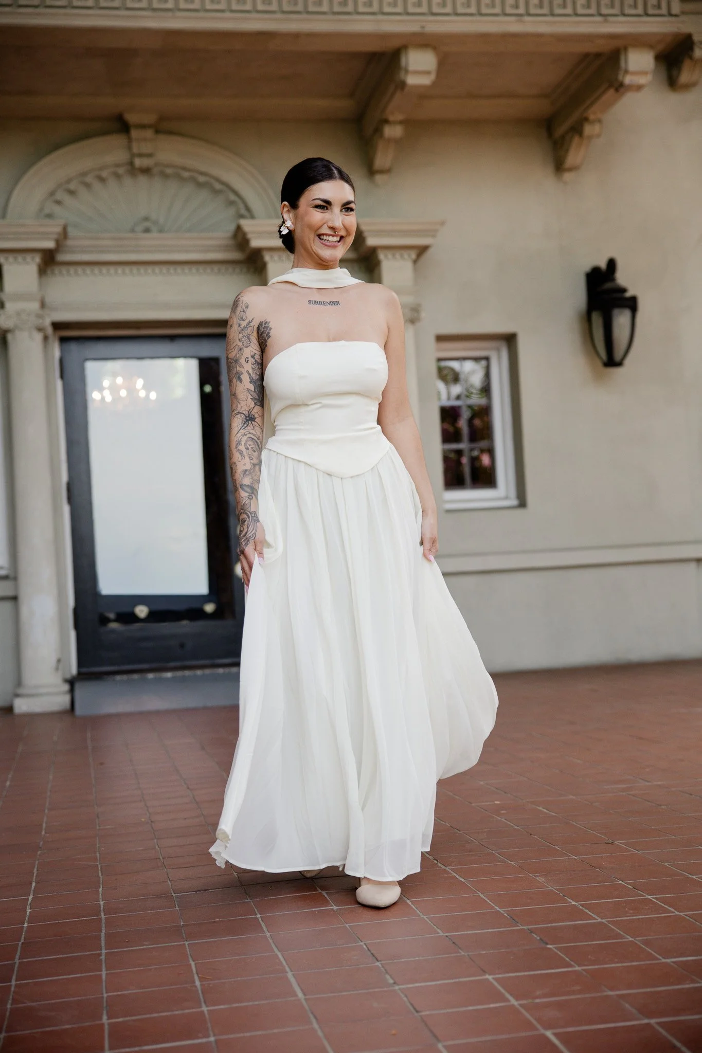A woman in a white strapless dress standing outdoors on a red-tile patio, smiling with visible tattoos on her left arm and a tattoo reading "SURRENDER" on her chest, in front of a building with a cream-colored wall and architectural details.