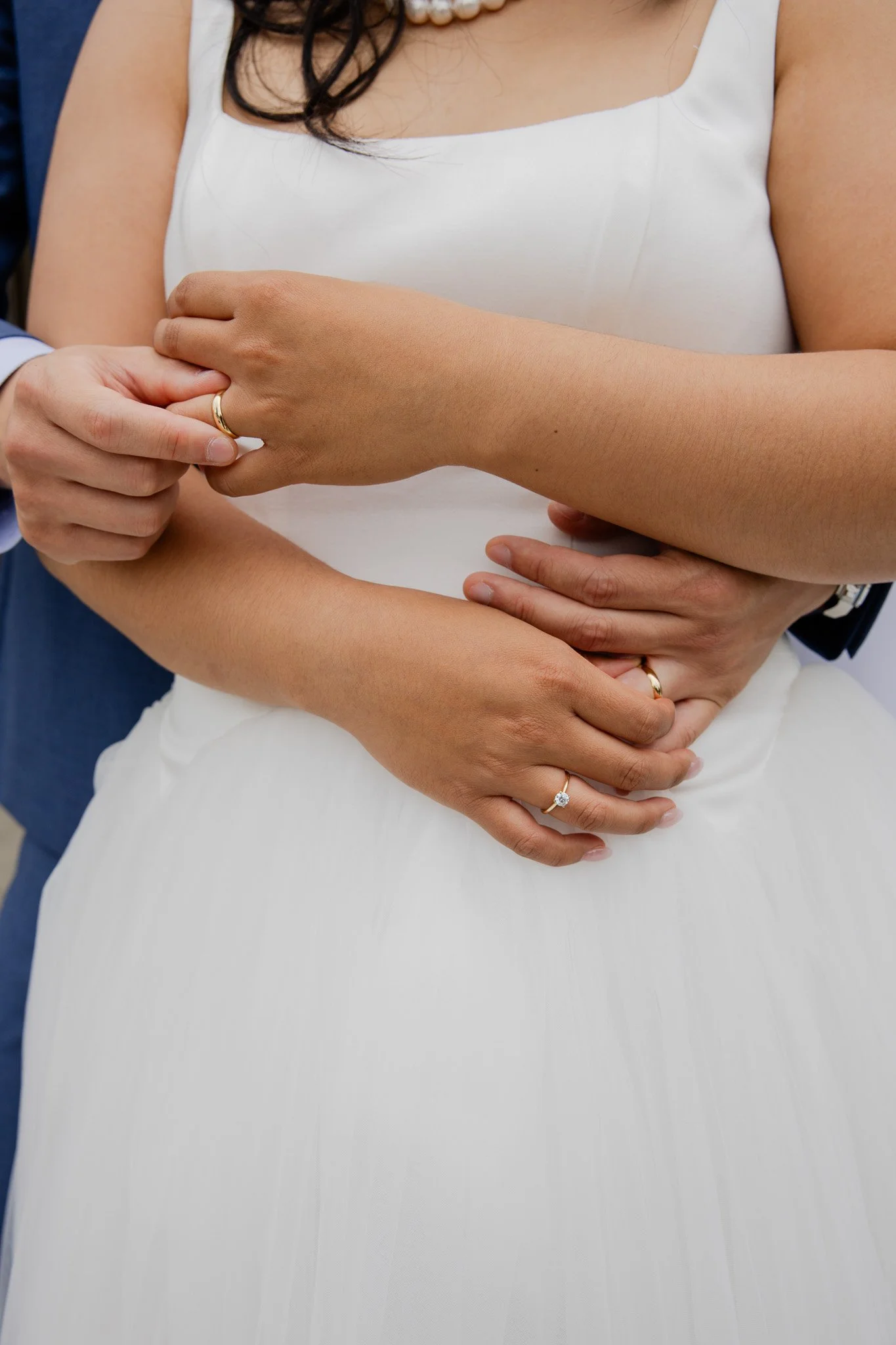 Close-up of a woman dressed in a white dress with a woman and a man holding her from behind in an embrace, highlighting her hands and jewelry.