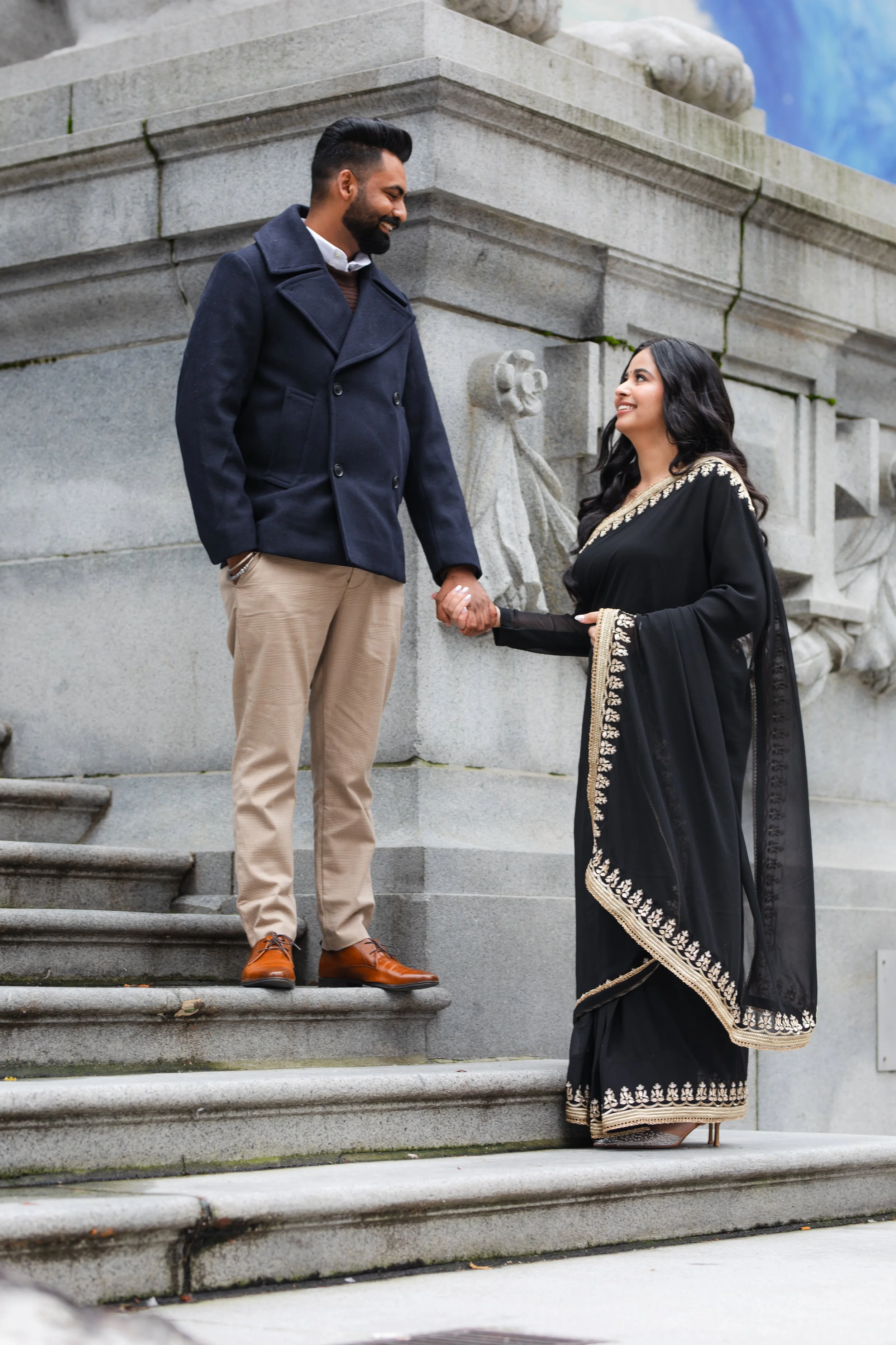 A man and woman holding hands on stone steps in front of a historical monument, smiling at each other. The man is wearing a navy coat and beige pants, the woman is dressed in a black saree with gold embroidery.