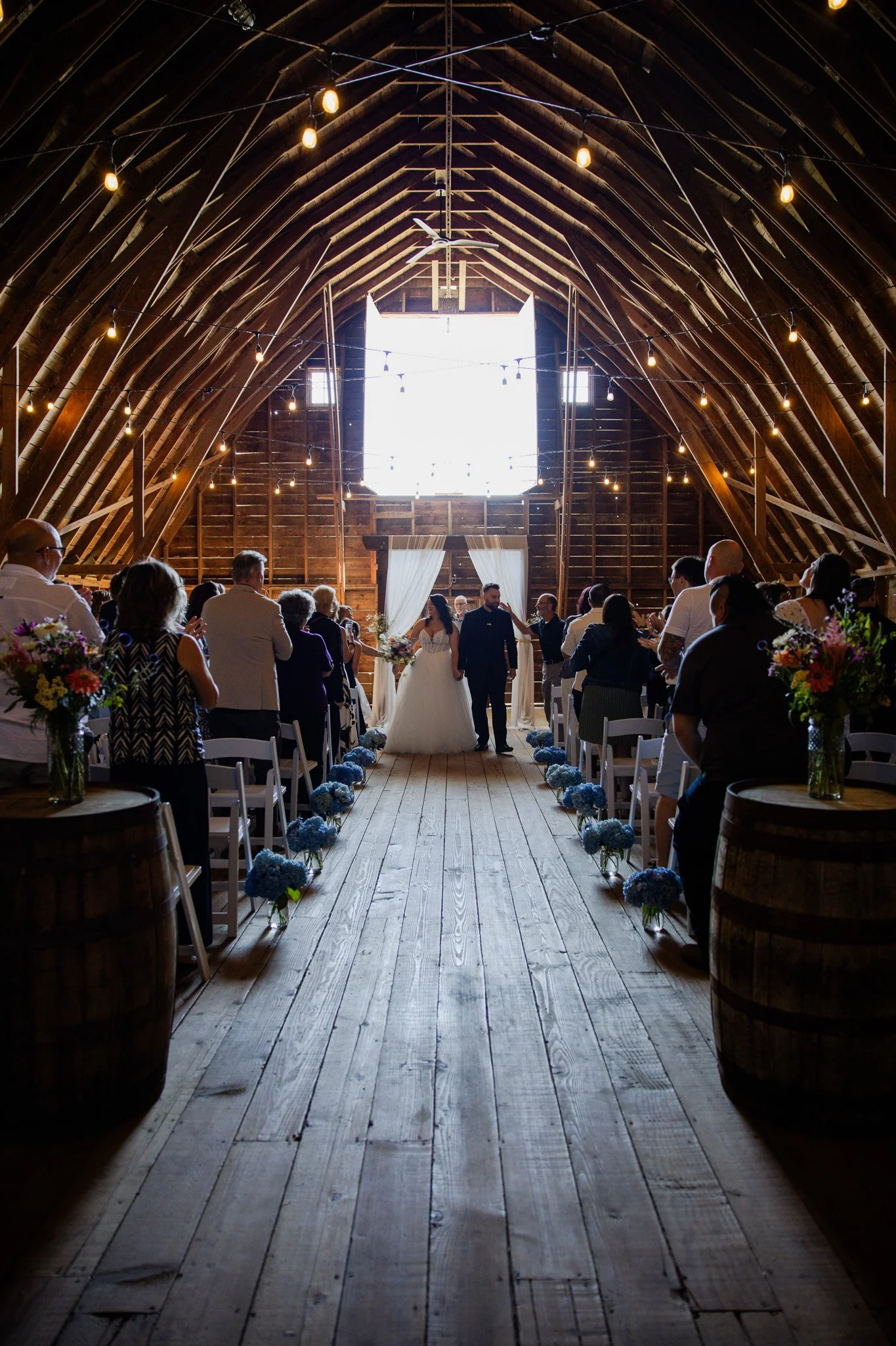 A wedding ceremony inside a rustic wooden barn with wooden beams and string lights hanging from the ceiling. The bride and groom are standing at the altar, surrounded by seated guests. The aisle is decorated with blue hydrangea flowers in small vases
