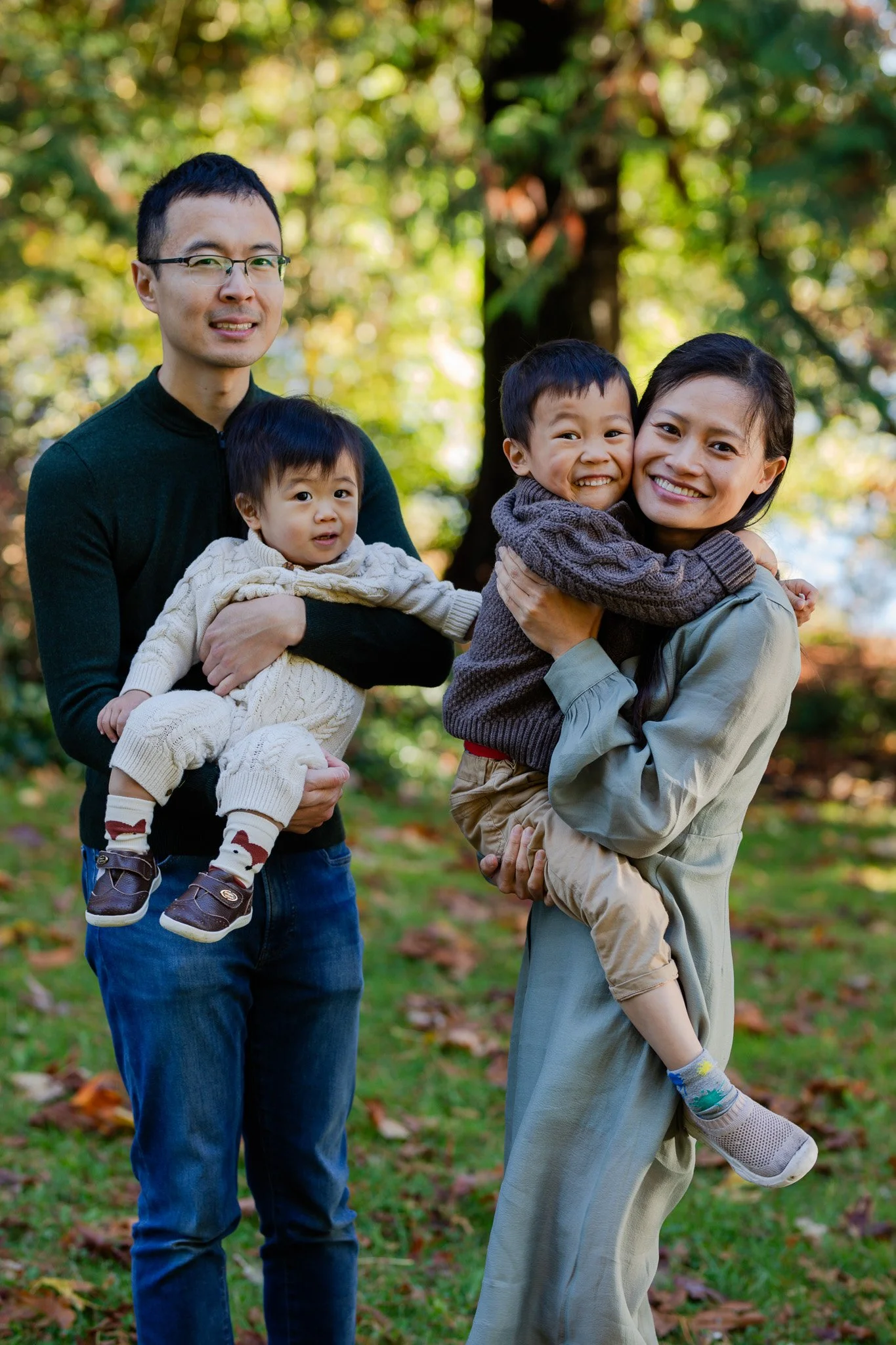 A family of four outdoors in a park on a fall day. The father is holding a young child, and the mother is holding an older child. They are all smiling and dressed warmly. Leaves are scattered on the ground, and trees with autumn foliage are in the ba