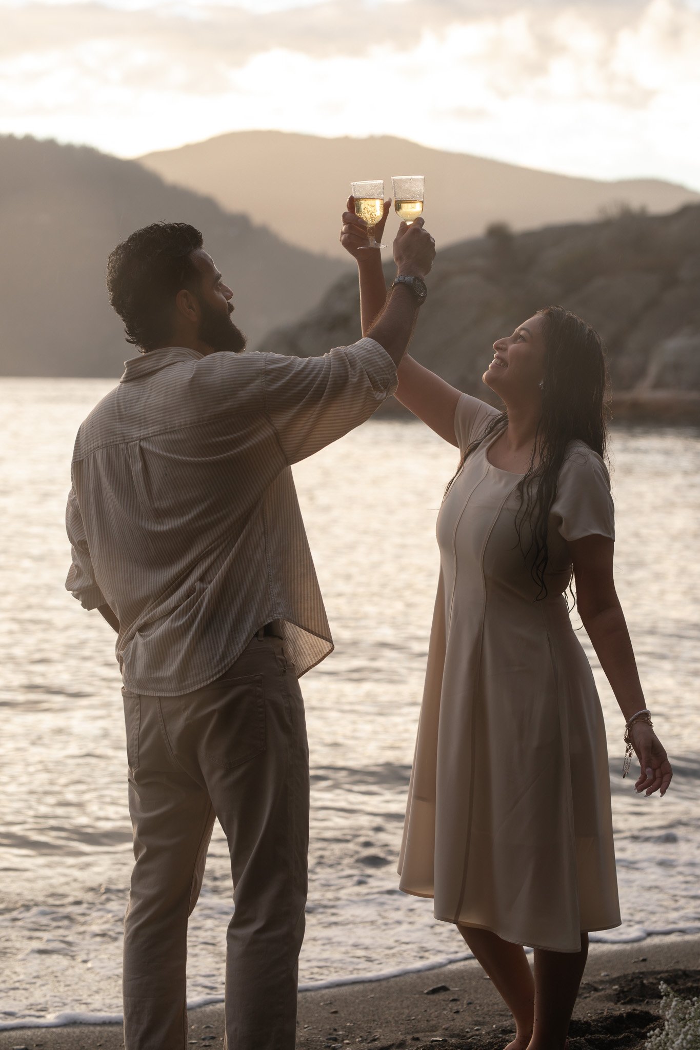 A man and woman raising champagne glasses in a toast on a beach at sunset, with mountains in the background.