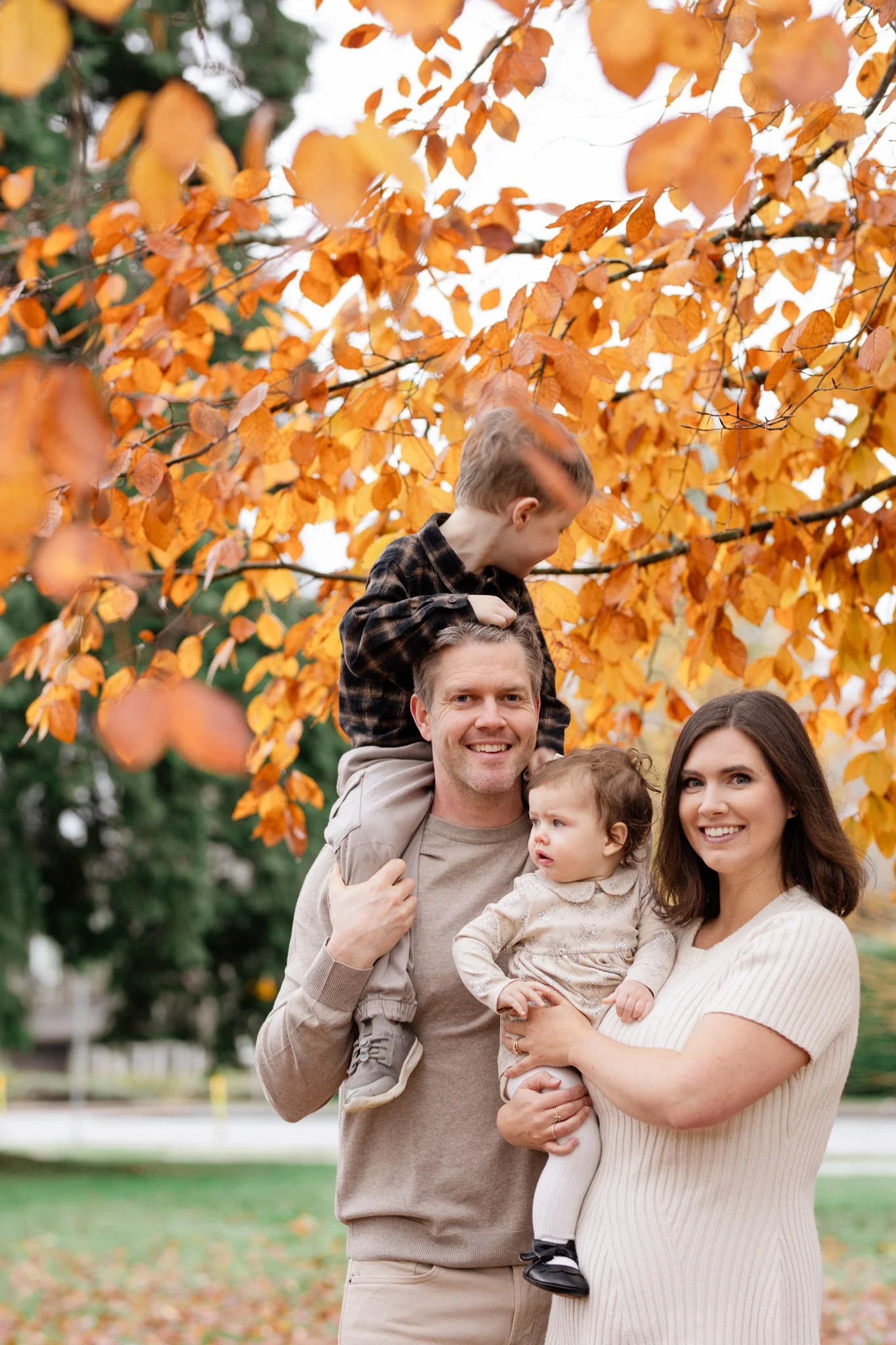 A family of four standing outdoors during fall, with orange and yellow leaves on a tree behind them. The father is smiling and carrying an older child on his shoulders, while the mother holds a younger child in her arms. Both children are looking aro