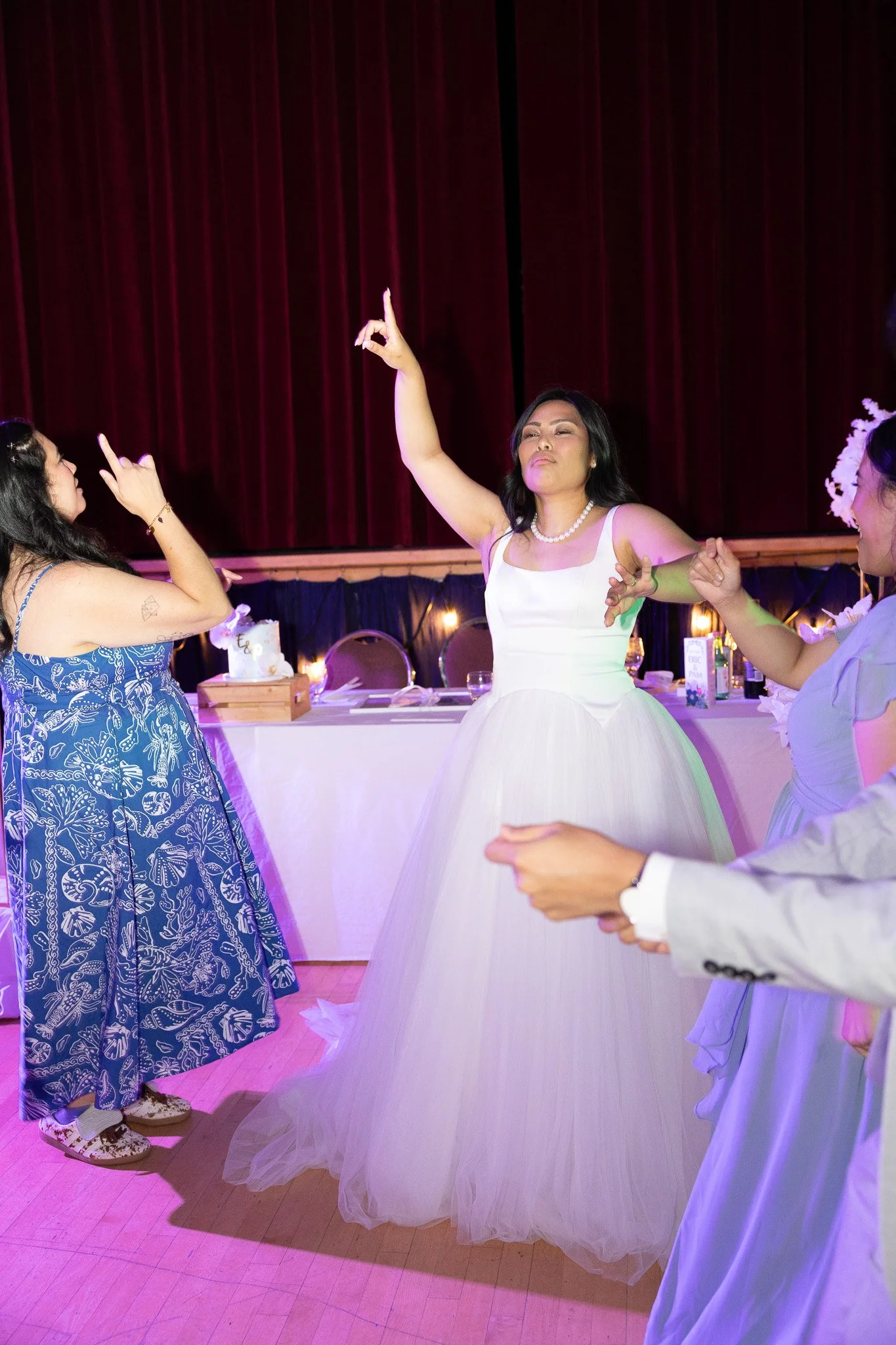 A woman in a white wedding dress dancing with her eyes closed inside a decorated reception hall with red curtains and tables.