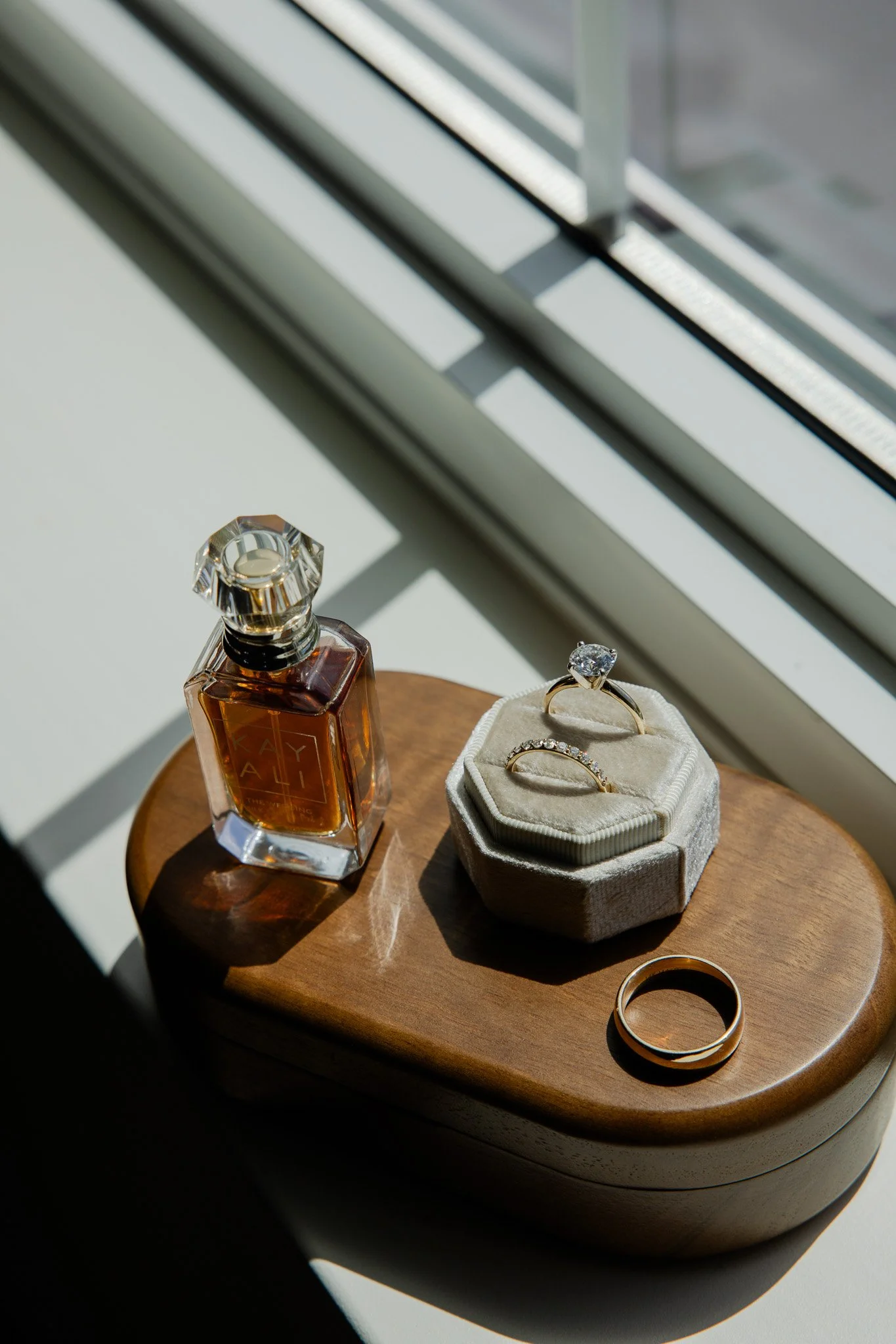 A wooden table with a bottle of perfume, two rings, and a jewelry box under a window with blinds, with sunlight casting shadows.