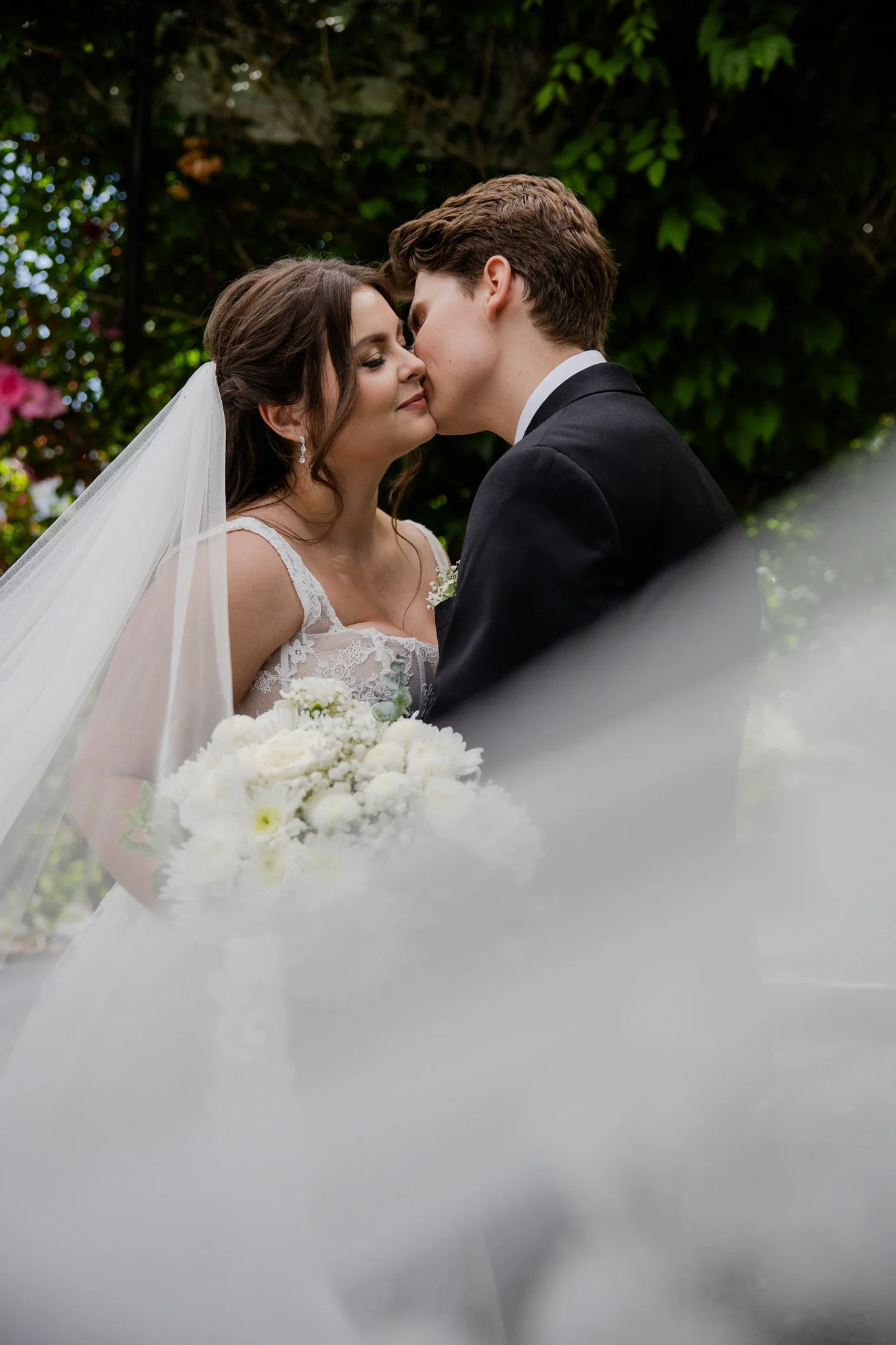 A bride and groom sharing a kiss outdoors during their wedding ceremony. The bride is wearing a white lace wedding dress and veil, holding a bouquet of white flowers. The groom is in a black tuxedo, with green foliage in the background.