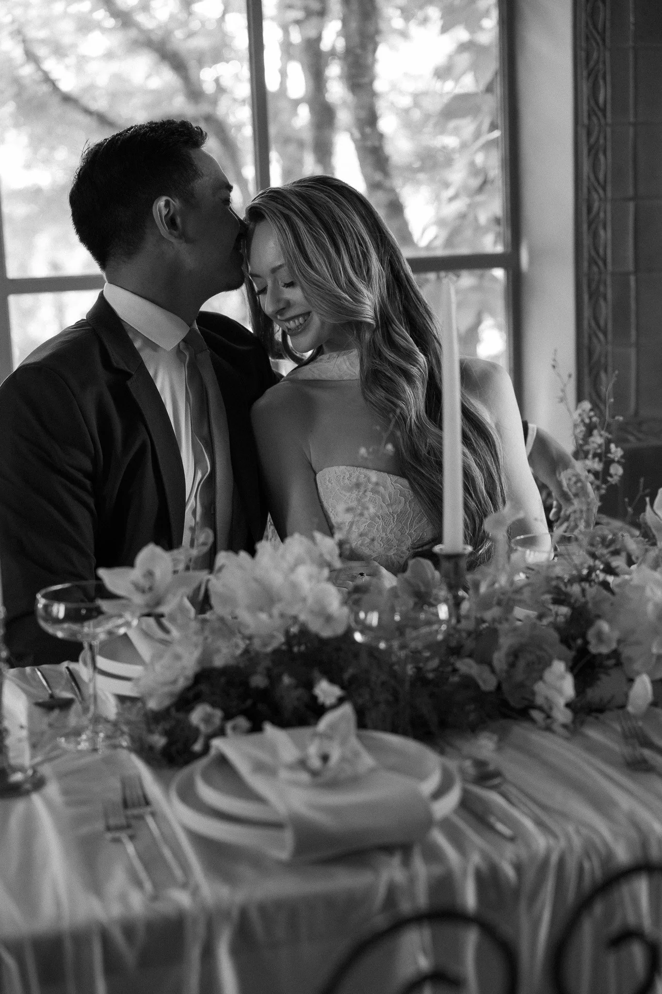 A couple sharing an intimate moment at a wedding or special event, with the man kissing the woman on the forehead and both smiling happily. There are flowers and candles on the table in front of them.