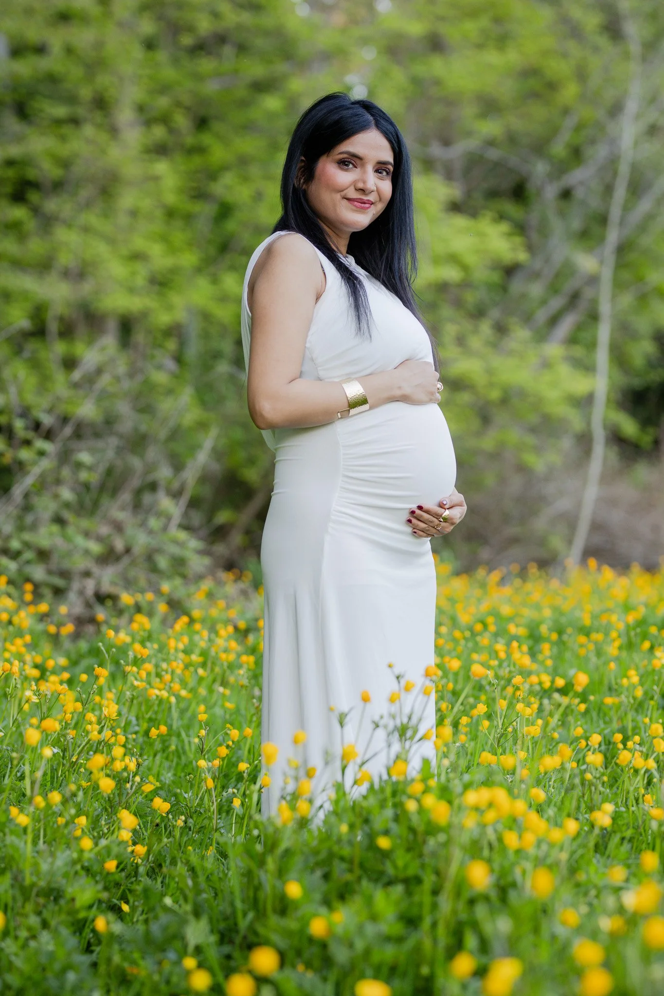 A pregnant woman with long dark hair wearing a white dress and gold jewelry standing in a field of yellow wildflowers with trees in the background.