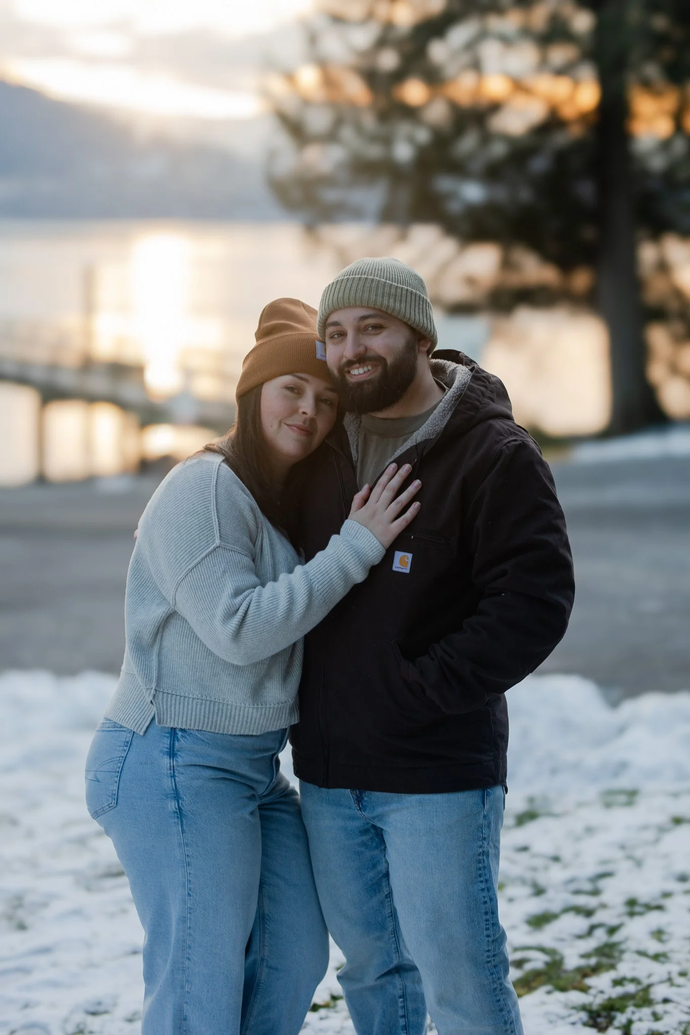A couple hugging outdoors on a snowy surface during sunset, with a large tree and a water body in the background.