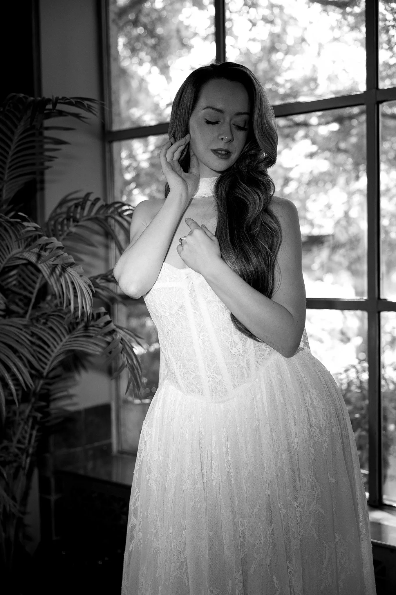 A woman in a wedding dress standing near large windows, with her eyes closed and hand touching her face, in black and white photograph.