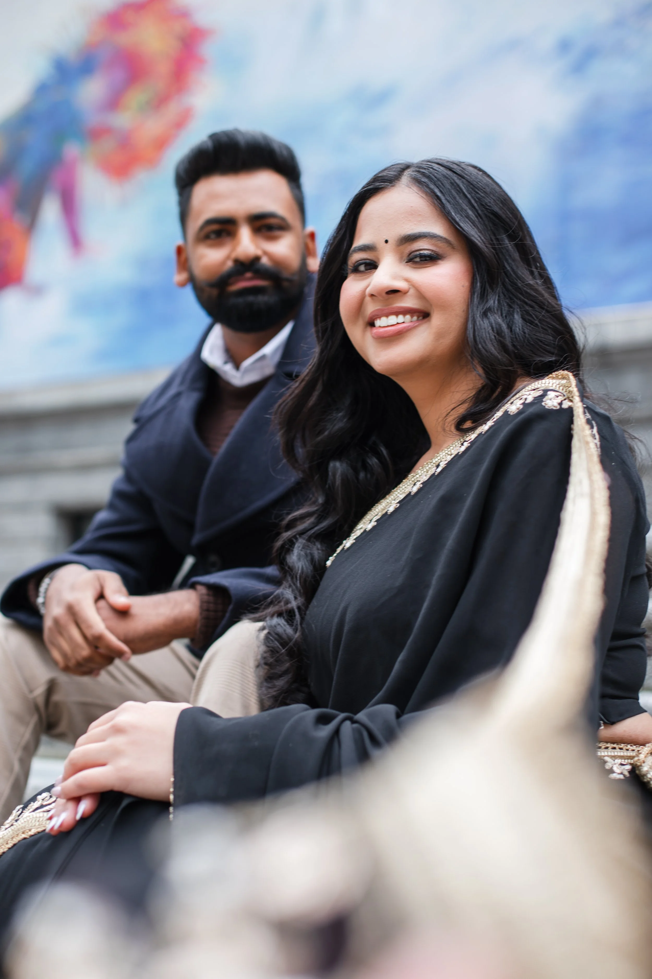 A smiling woman in a black traditional Indian dress with gold embellishments sitting outdoors, with a man in a dark coat and beige pants sitting beside her, both against a blurred background of an artistic mural.