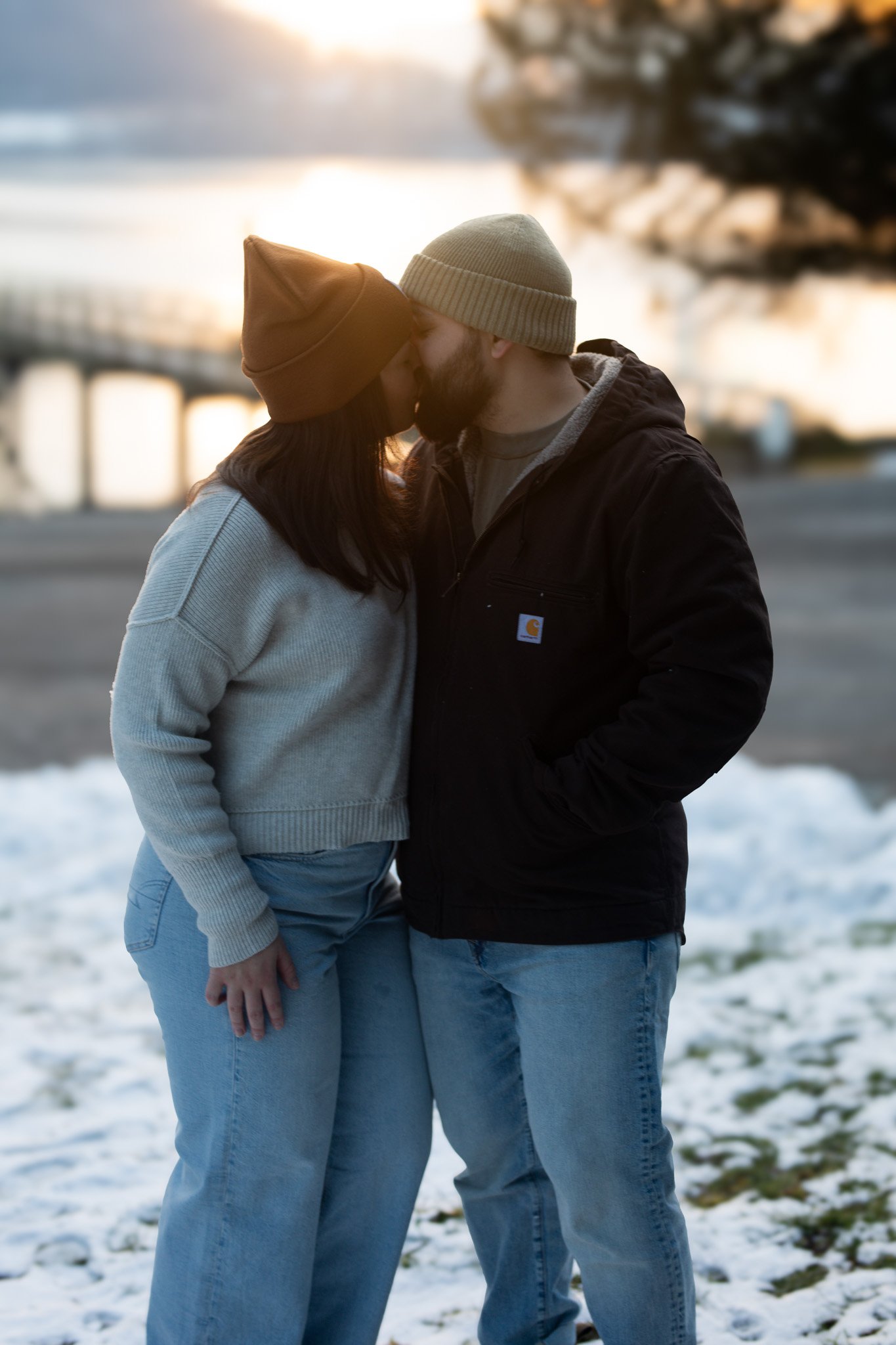 A couple standing close together, kissing on a beach with snow on the ground during sunset, both wearing casual winter clothing.