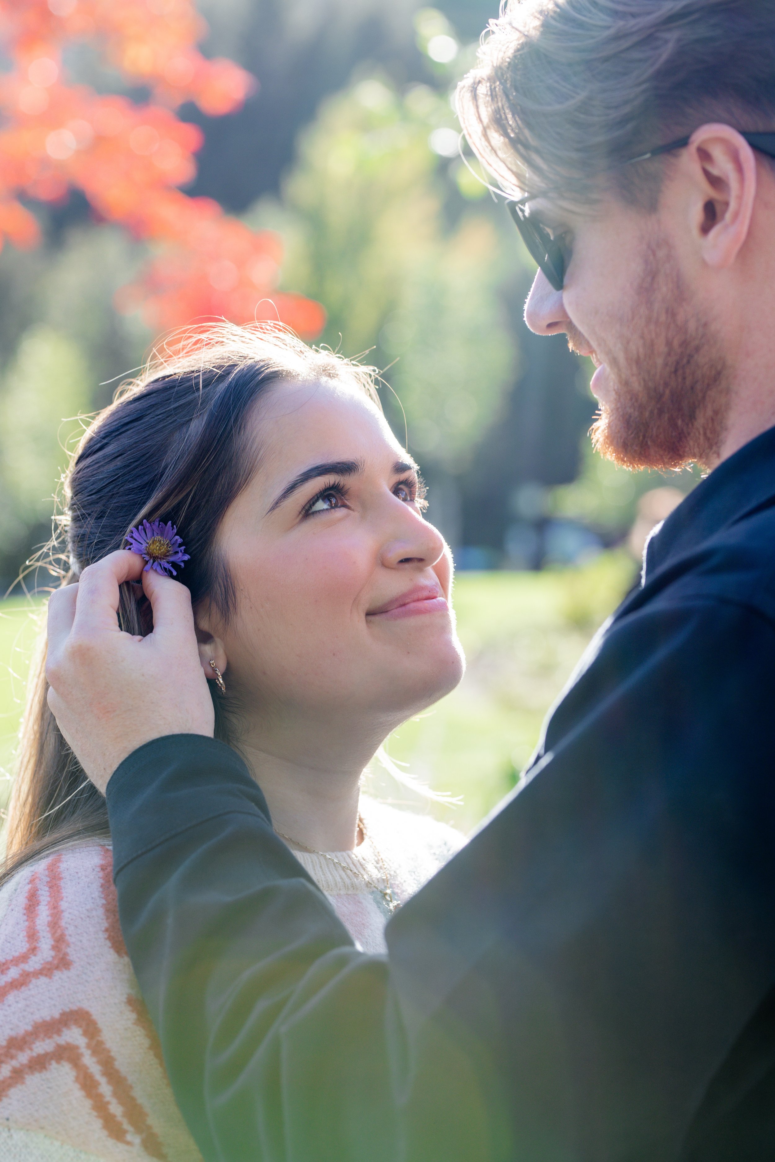 A young woman with dark hair and a purple flower in her ear looks up at a young man with glasses and a beard, as he gently touches her face outdoors in a park during fall.