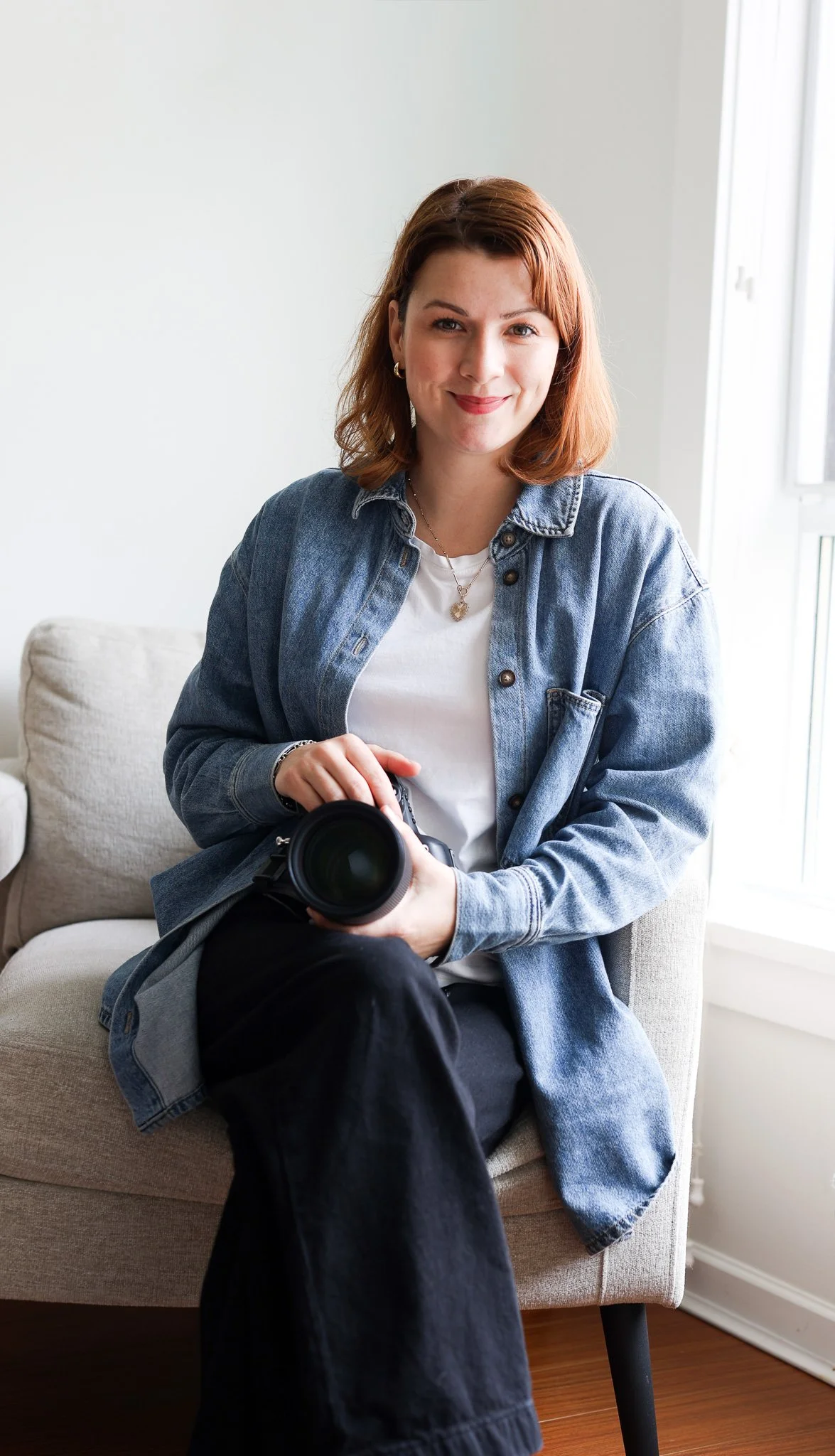 A young woman with red hair and a denim jacket sitting on a beige couch, holding a camera, smiling at the camera in a bright room with white walls and a window.