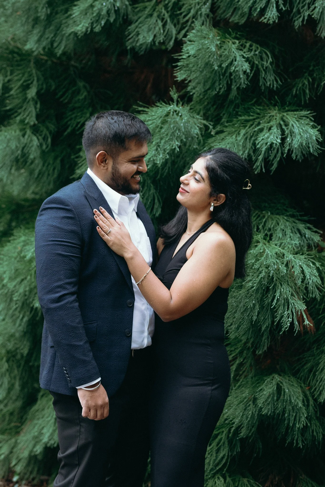 A couple dressed formally embracing outdoors in front of green pine tree branches, sharing a loving moment.