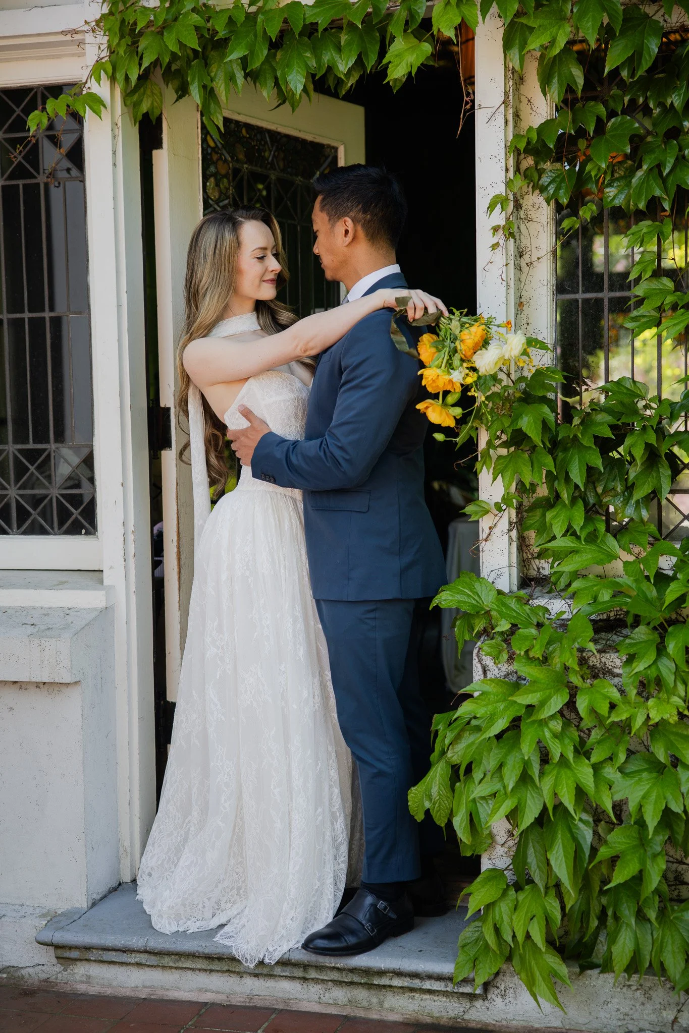 A bride and groom are standing on a porch, looking at each other lovingly. The bride is wearing a white lace wedding gown and has long blonde hair. The groom is dressed in a dark blue suit with a tie. The bride is holding a bouquet of yellow and whit
