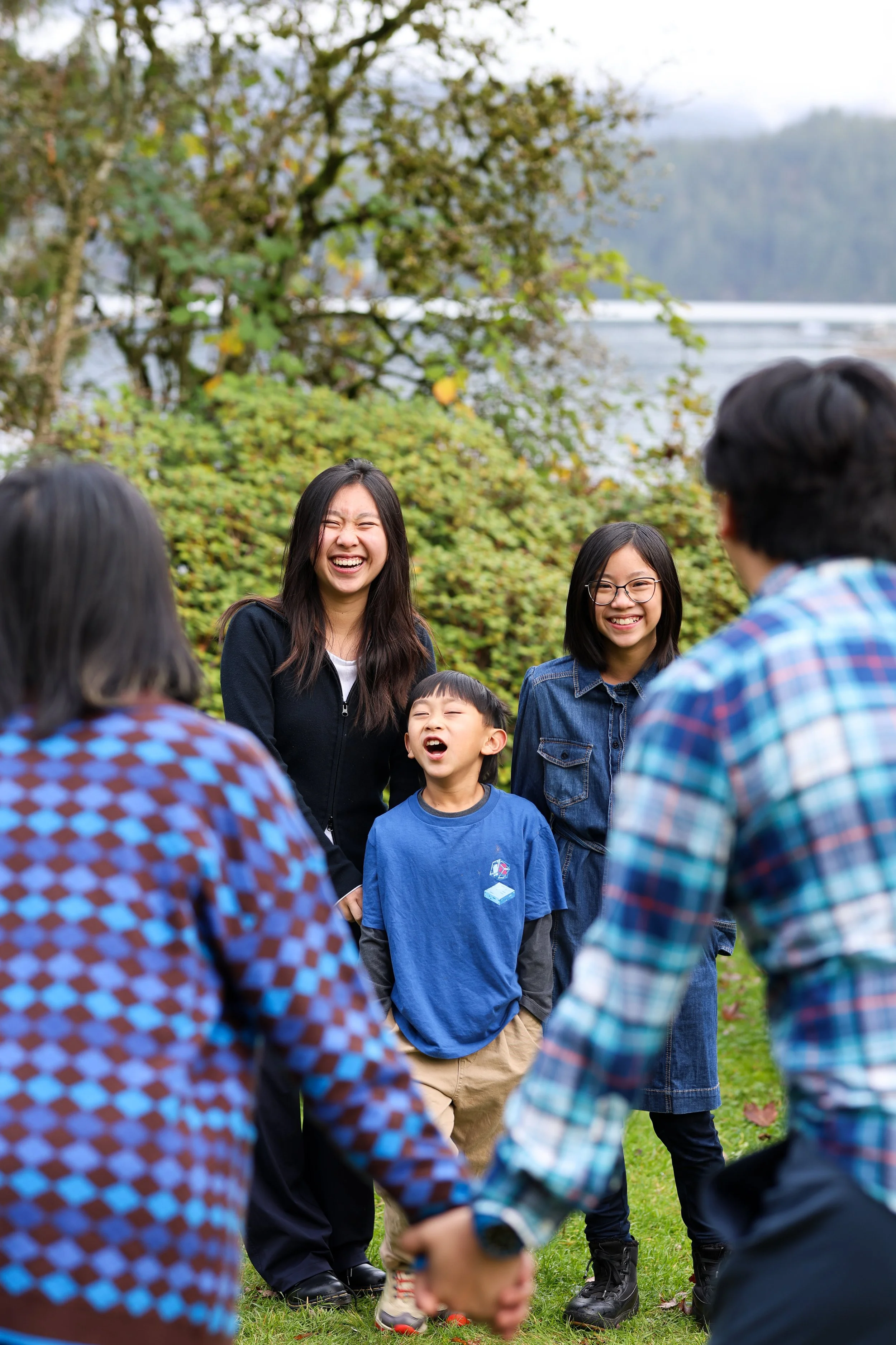 Group of diverse kids and teenagers holding hands and laughing outdoors on a grassy area with trees in the background.