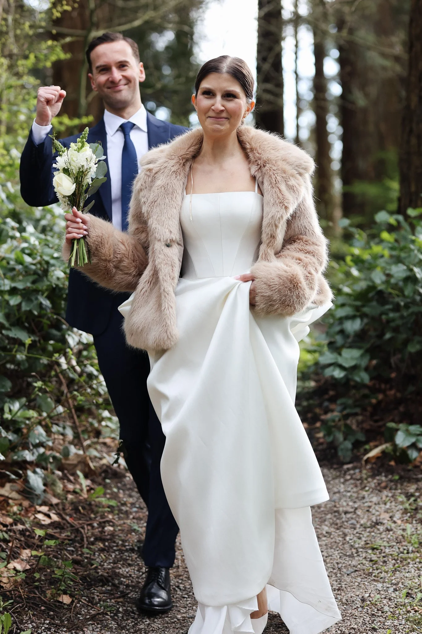 A bride in a white wedding dress and a fur coat holding a bouquet of white flowers stands in front of a groom in a suit and tie, with his fist raised, in a forested area.