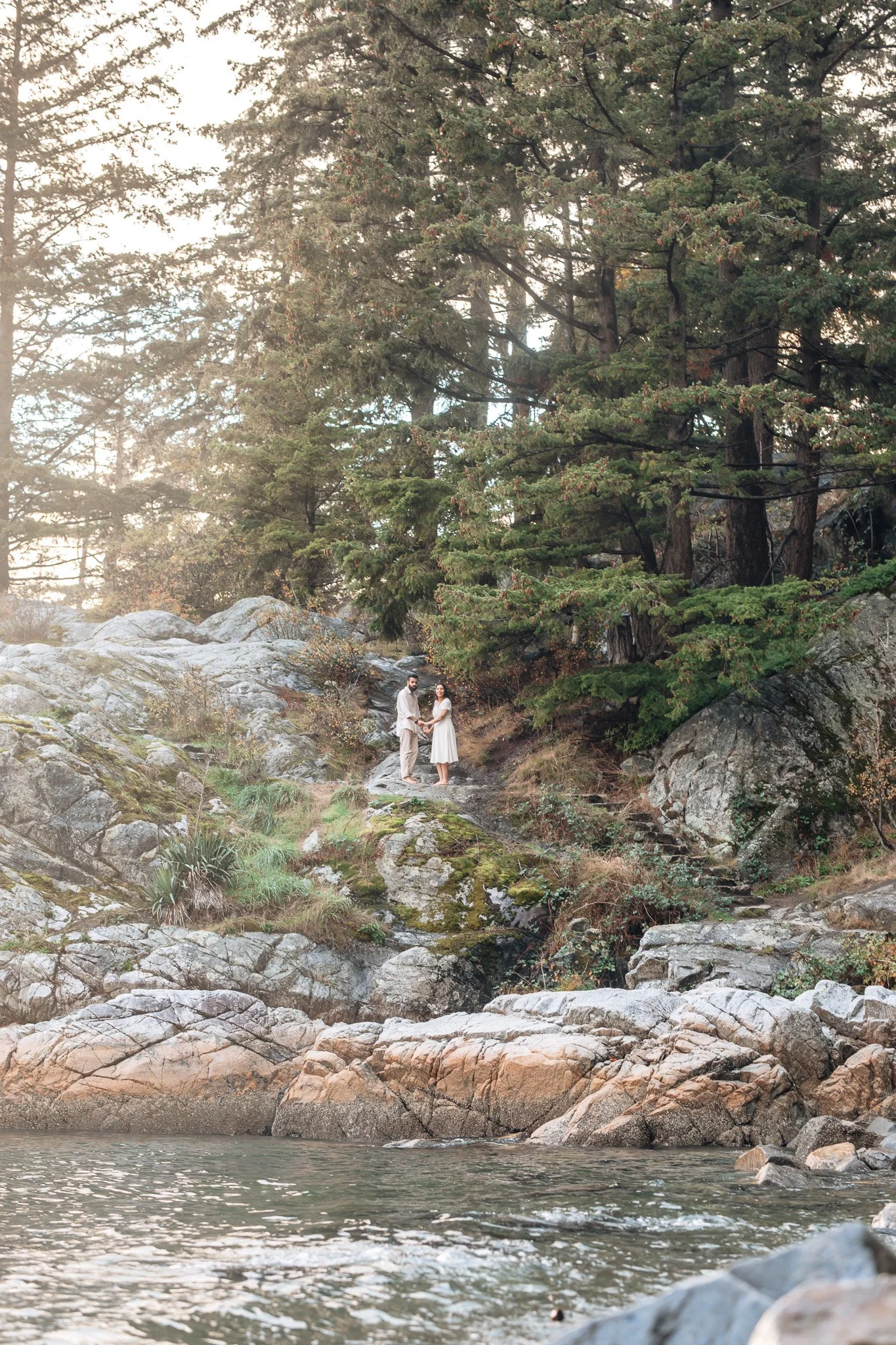 A couple dressed in light-colored clothing holding hands on rocky terrain near water, surrounded by tall trees in a forest setting.